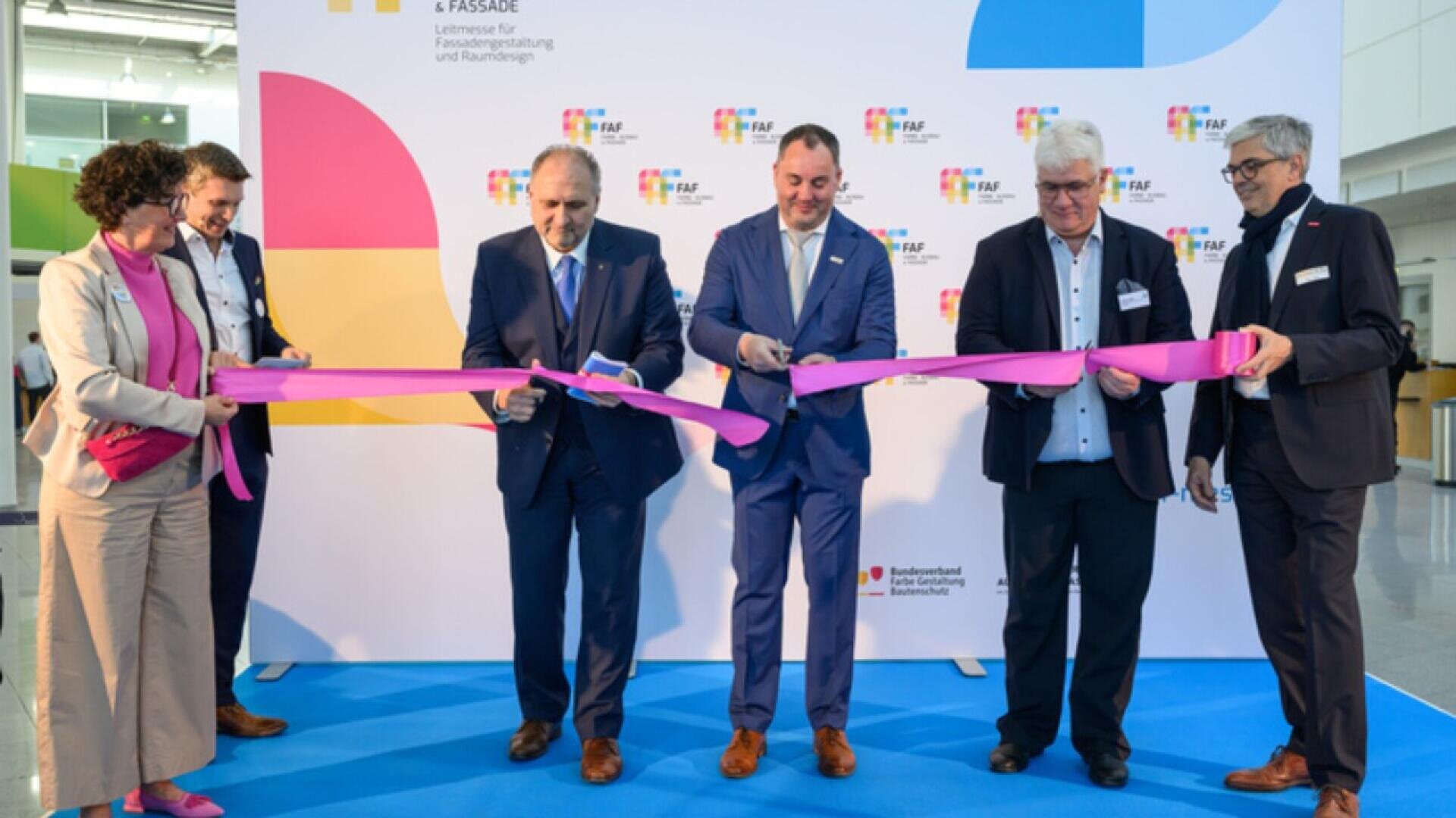Six people in business attire cut a long pink ribbon together at an event, standing on a blue carpet in front of a white backdrop with colorful logos and text.