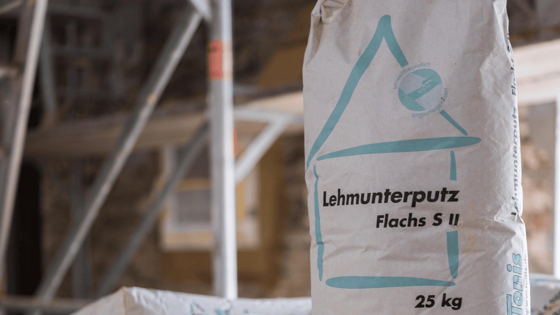 Nahaufnahme eines 25-kg-Sacks mit der Aufschrift Lehmunterputz Flachs S II und einem aufgedruckten blauen Hauslogo vor einem unscharfen Baustellenhintergrund mit Gerüsten und einer Steinmauer.