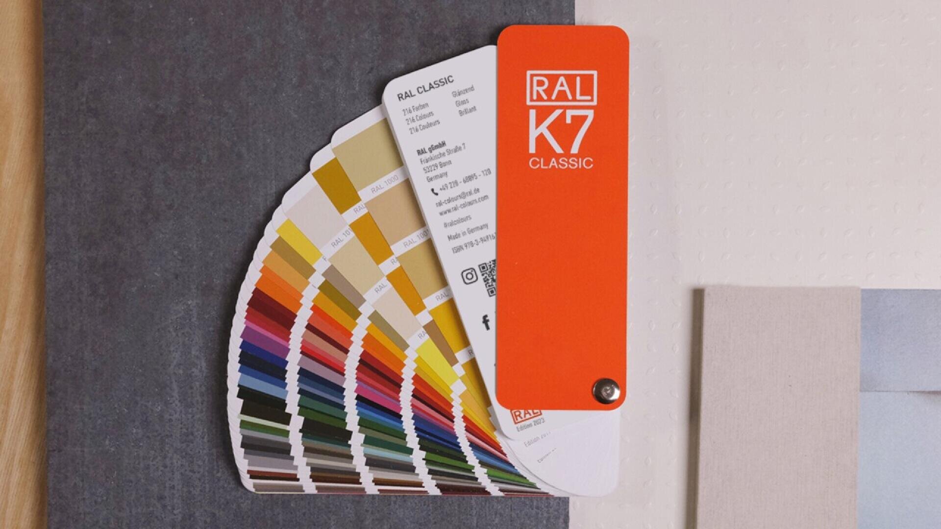 A RAL K7 Classic color fan deck is spread open, displaying a wide range of color swatches. The top card is bright orange with white text. The deck rests on a dark surface with beige and light gray surrounding it.