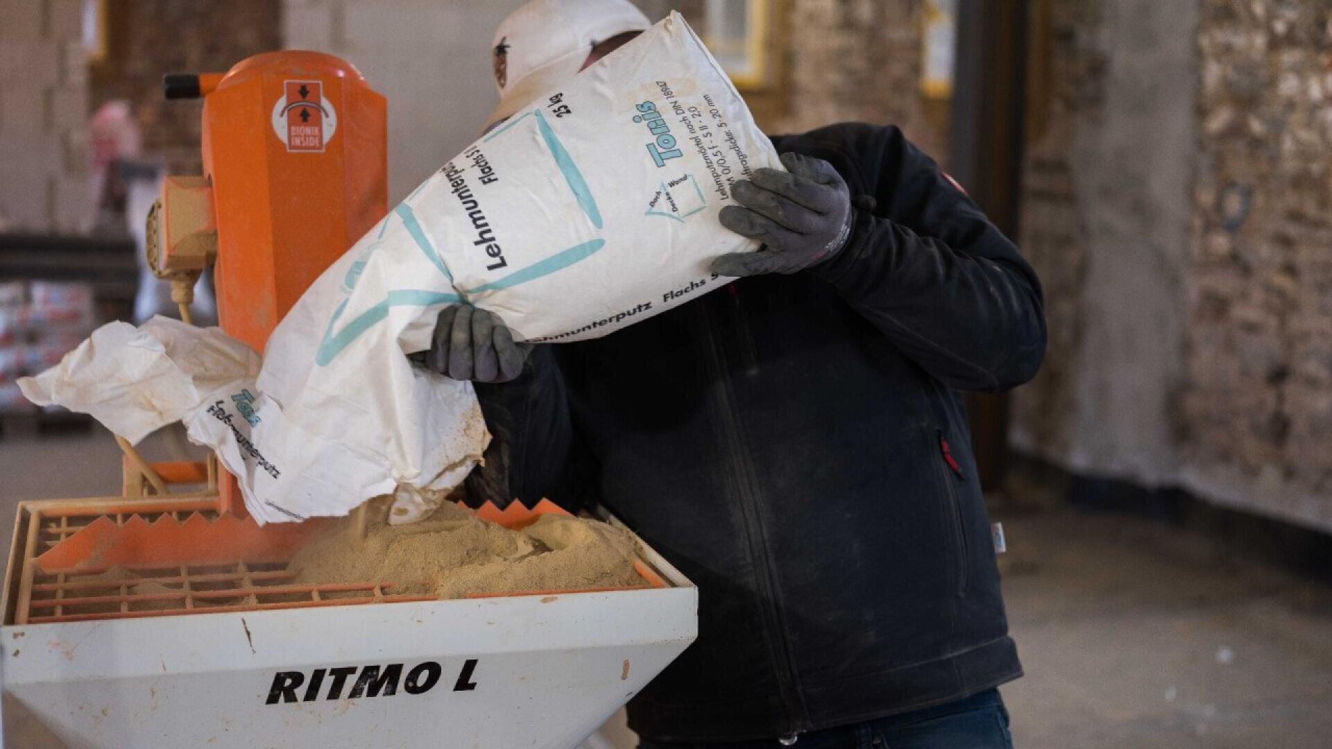 A person in work gloves and a hard hat pours a bag of material into an orange Ritmo L machine inside a construction site with exposed brick walls.
