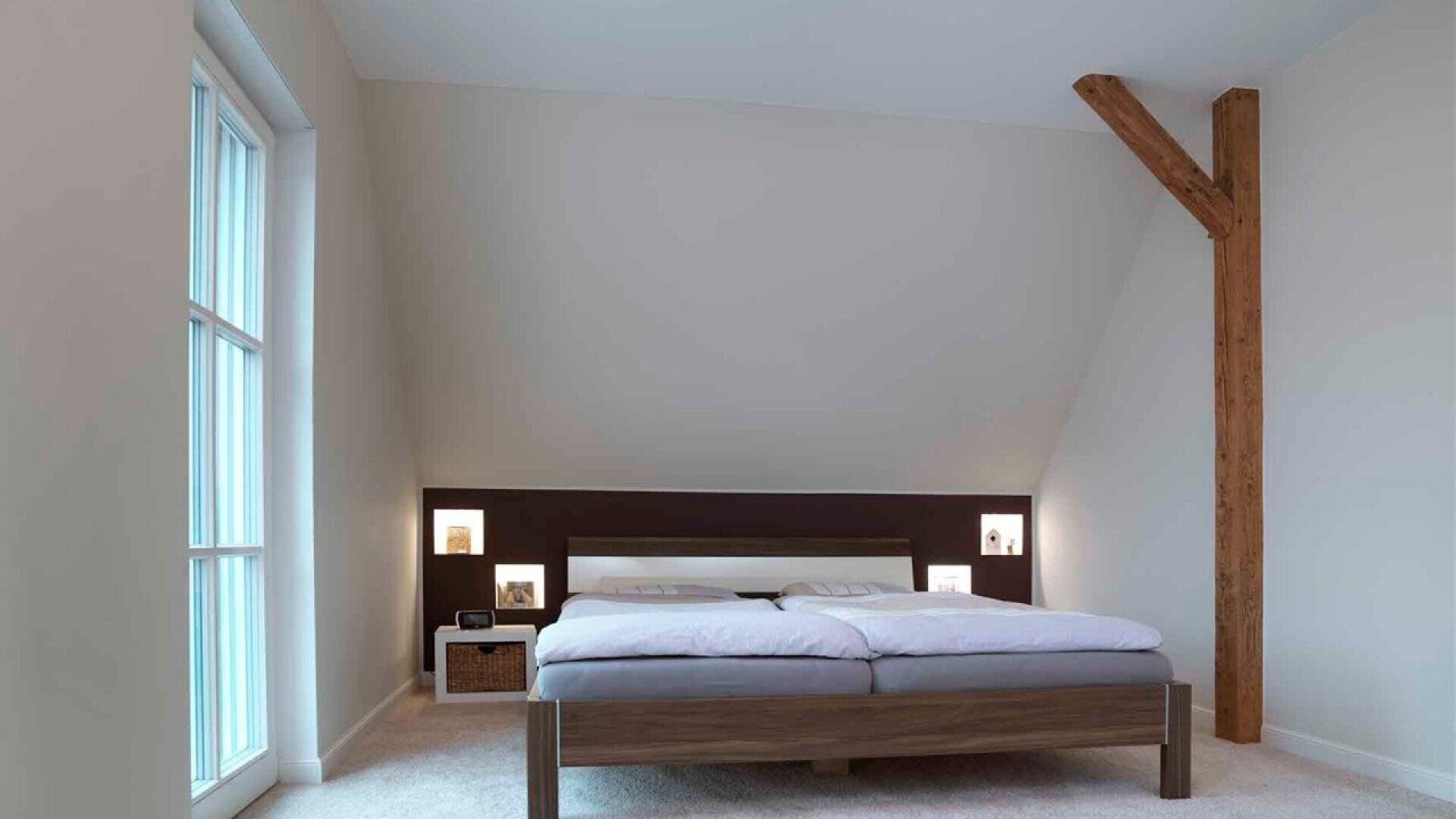 Minimalist bedroom with a wooden bed, white bedding, and built-in headboard shelves with lights. Large window on the left and an exposed wooden beam on the right wall. Neutral walls and carpeted floor.