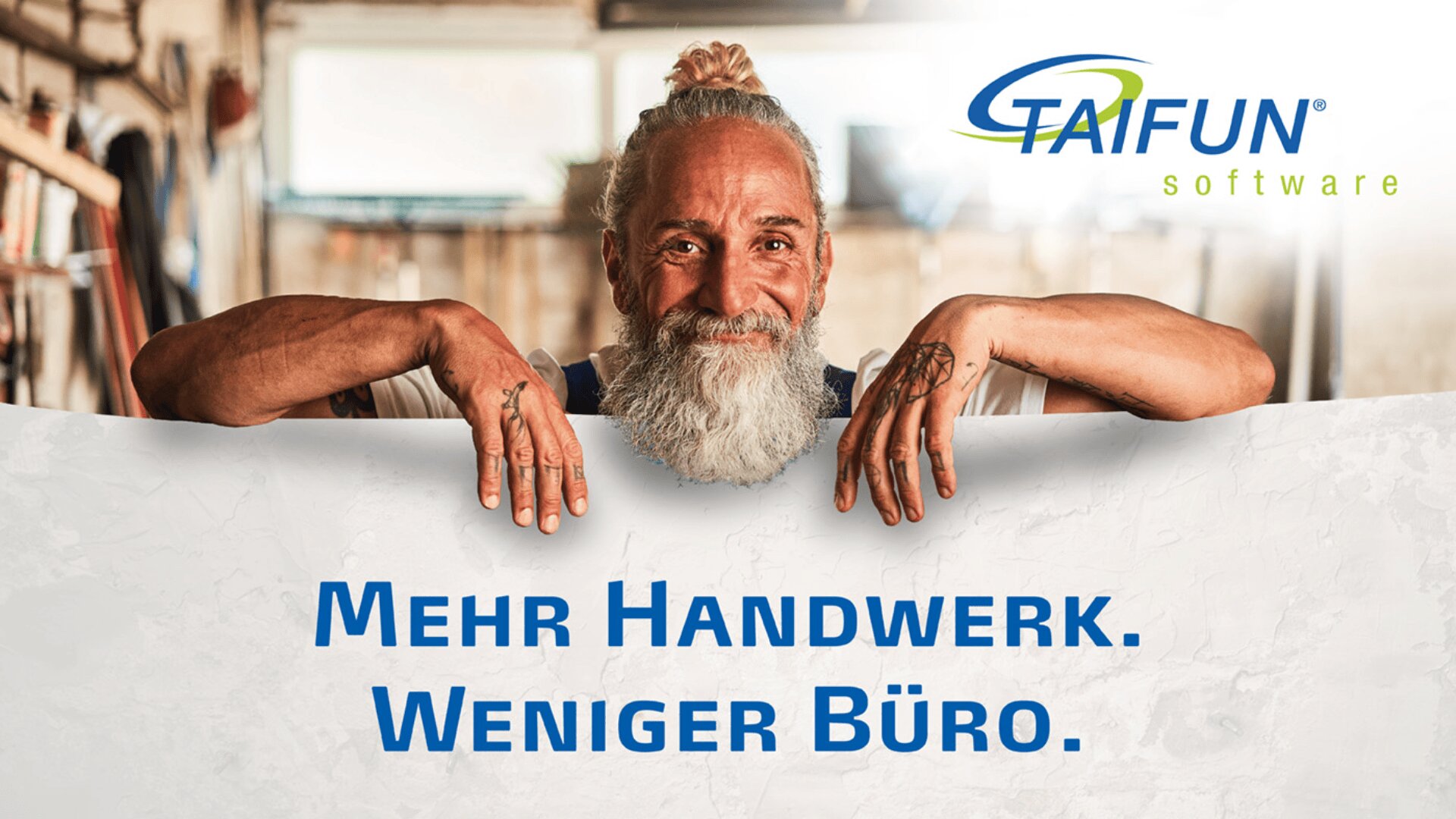 Ein lächelnder Mann mit langem weißen Bart hält ein Schild mit der Aufschrift „Mehr Handwerk. Weniger Büro“. Oben rechts befindet sich das Logo der TAIFUN-Software. Im Hintergrund ist eine Werkstattumgebung zu sehen.