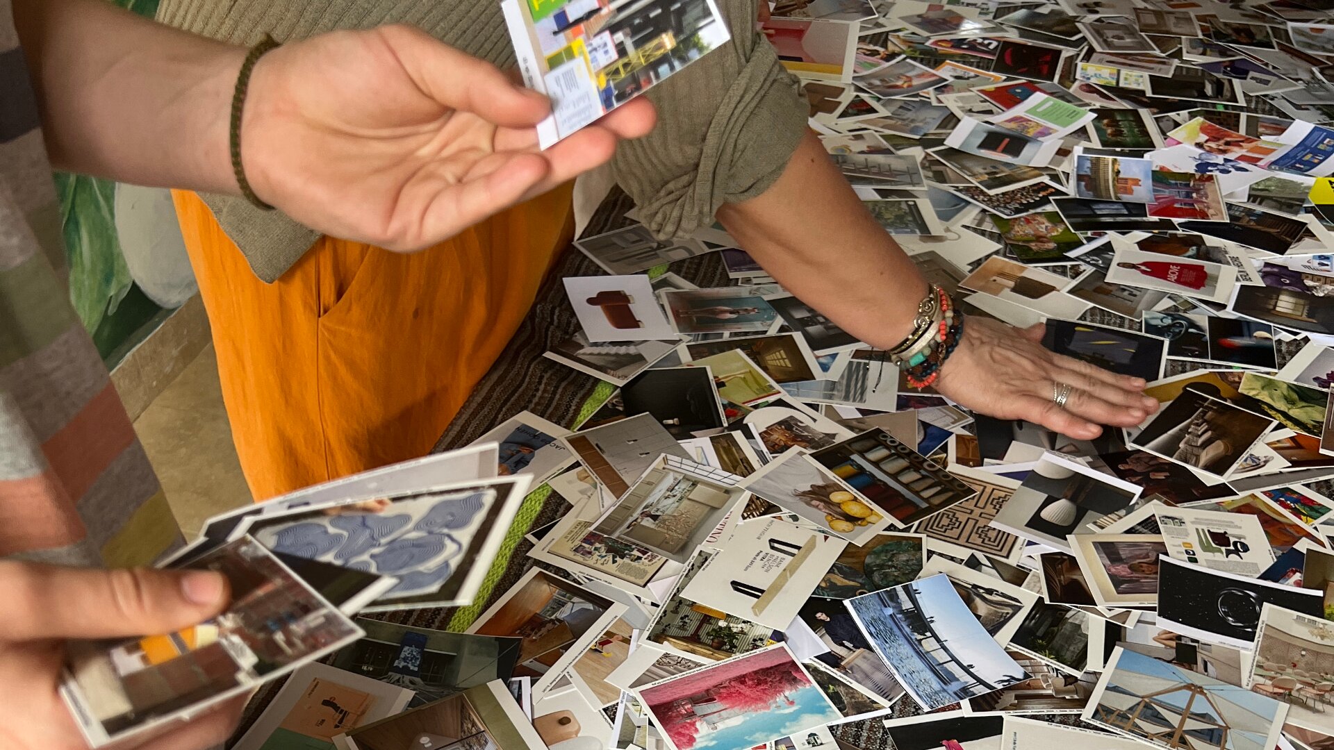 Zwei Personen sortieren eine große Sammlung kleiner, bunter Fotos, die auf einem Tisch ausgebreitet sind. Eine Person hält mehrere Fotos, während die andere die verstreuten Bilder ordnet oder auswählt.