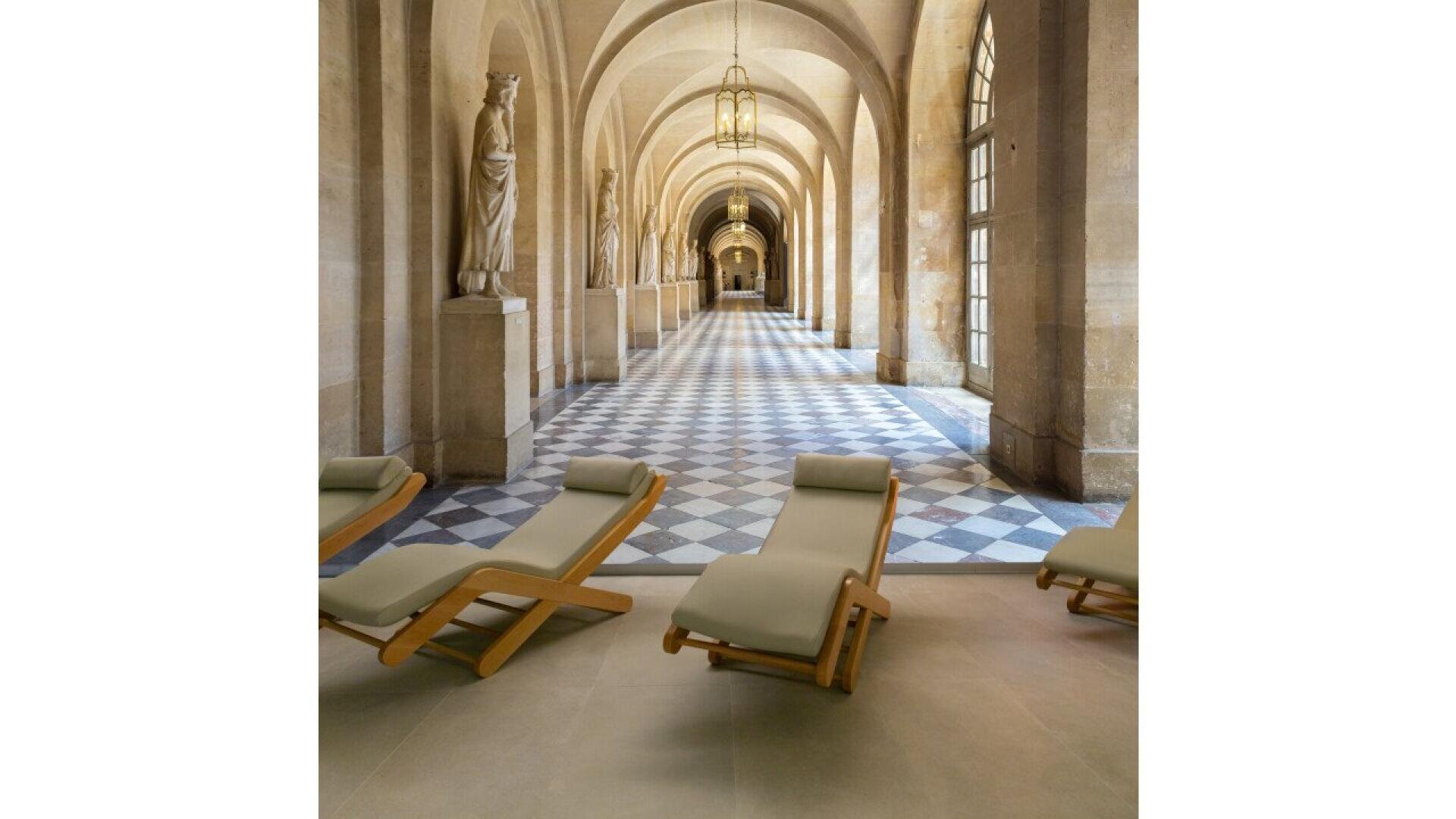 A grand hallway with arched ceilings, statues along the walls, black-and-white checkered floor tiles, and modern wooden lounge chairs with light cushions in the foreground.