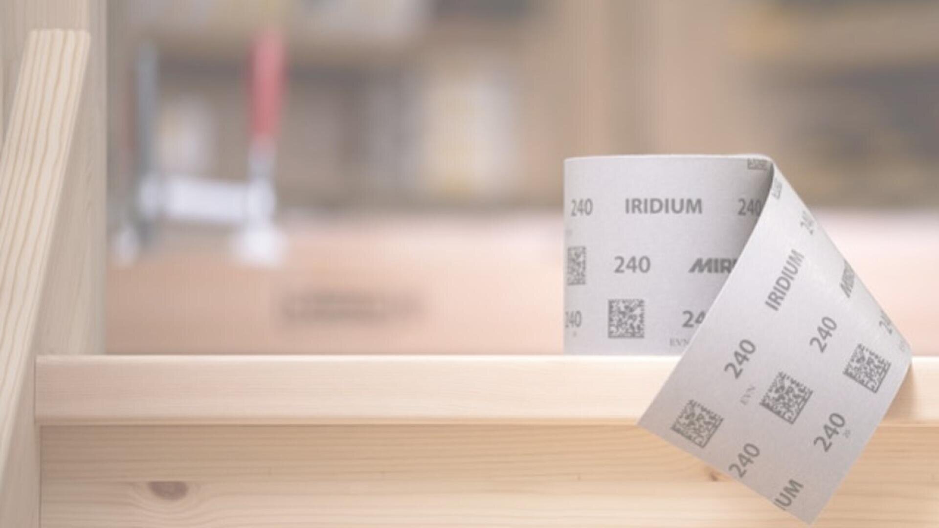 Eine Rolle Schleifpapier mit der Aufschrift IRIDIUM und 240 liegt auf einer hellen Holzoberfläche, im Hintergrund sind Regale und verschwommene Werkzeuge zu sehen.