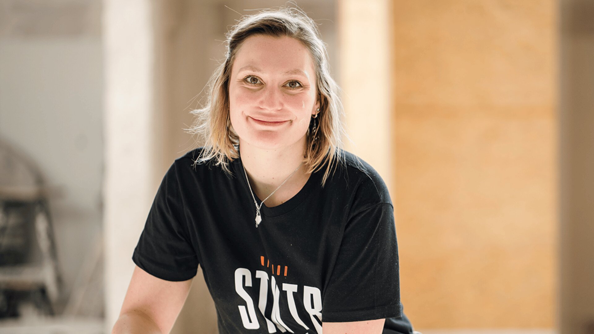 Eine Frau mit schulterlangem blondem Haar lächelt in die Kamera. Sie trägt ein schwarzes T-Shirt mit weiß-orangefarbenem Text und steht in einem sanft beleuchteten Raum mit neutralen Wandfarben.