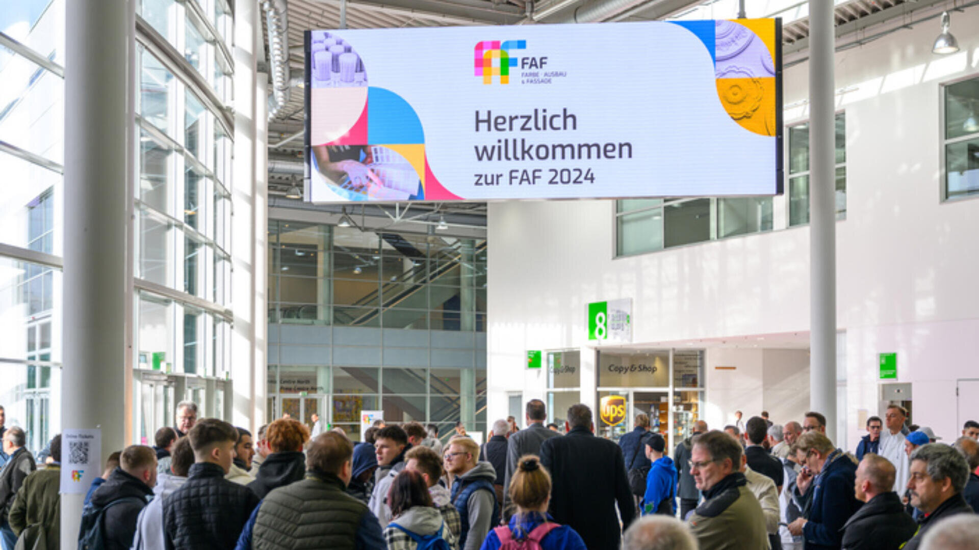 Eine Menschenmenge versammelt sich in einer hellen Ausstellungshalle unter einem großen Schild mit der Aufschrift „Herzlich willkommen zur FAF 2024“. Sonnenlicht strömt durch hohe Fenster.