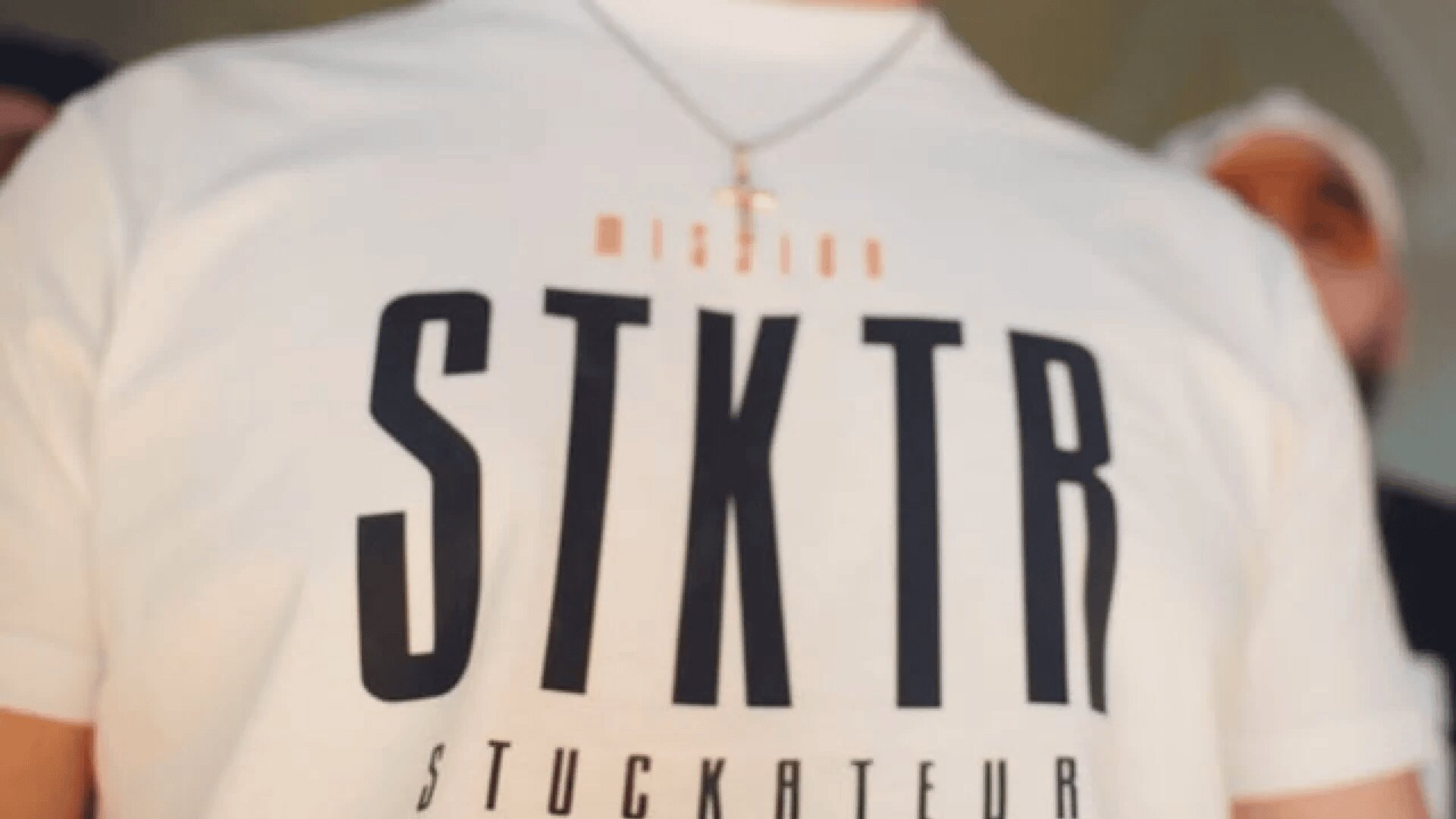 Eine Person trägt ein weißes T-Shirt mit den fettgedruckten schwarzen Buchstaben „STKTR“ und dem kleineren Text „STUCKATEUR“. Darüber steht in kleinerer Schrift das Wort „MISSION“. Die Person trägt außerdem eine silberne Kette mit einem Kreuzanhänger.