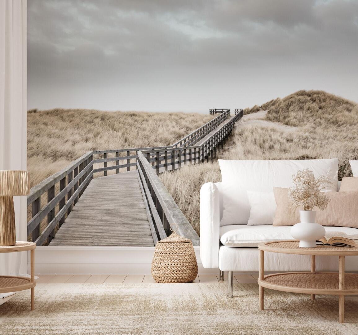 Ein gemütliches Wohnzimmer mit neutralem Dekor verfügt über ein großes Wandbild mit einem Holzsteg, der unter einem bewölkten Himmel durch grasbewachsene Dünen führt und so die Illusion von Tiefe und einer Landschaft im Freien erzeugt.