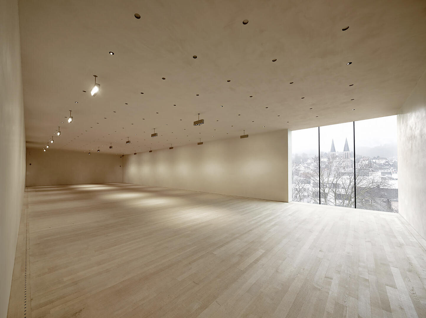 Ein großer, leerer, minimalistischer Raum mit hellem Holzboden, beigen Wänden und einer Decke mit Einbauleuchten. An einem Ende des Raumes befindet sich ein großes Fenster mit Blick auf eine Stadtlandschaft mit Kirchtürmen.
