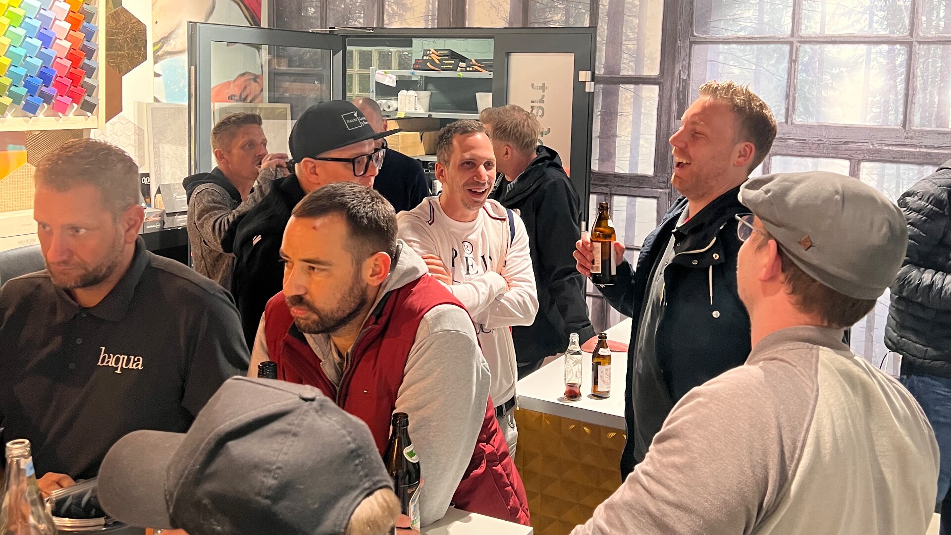 Eine Gruppe Männer trifft sich in einer hell erleuchteten Bar, plaudert, lacht und hält Getränke in der Hand. Flaschen stehen auf der Theke, einige Männer tragen Hüte und Jacken. Im Hintergrund sind ein Kühlschrank und ein großes Fenster zu sehen.