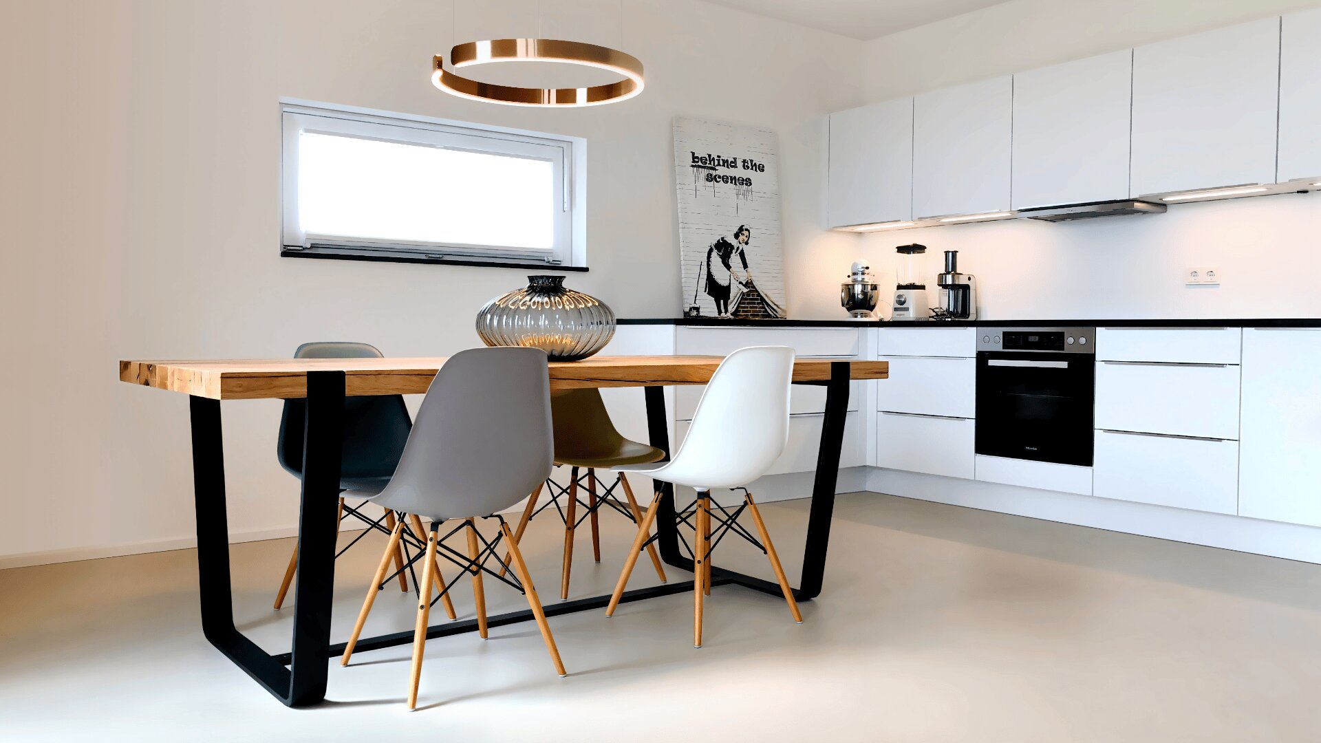 Moderne Küche und Essbereich mit einem Holztisch, vier verschiedenfarbigen Stühlen, einer runden Glasvase, einer runden Deckenleuchte, weißen Schränken, Einbaugeräten und einem Wandkunstwerk mit einer Frau.