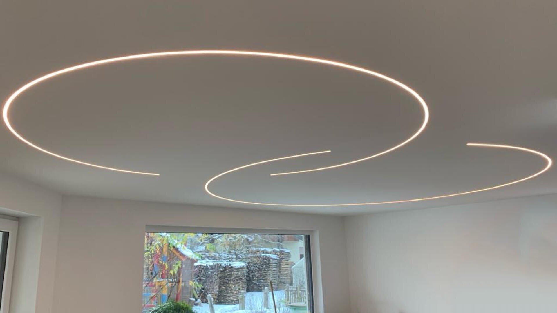 Eine weiße Decke ist mit modernen, kreisförmigen LED-Lichtstreifen versehen, die große leuchtende Ringe über einem hellen Raum mit einem Fenster und weißen Wänden bilden.