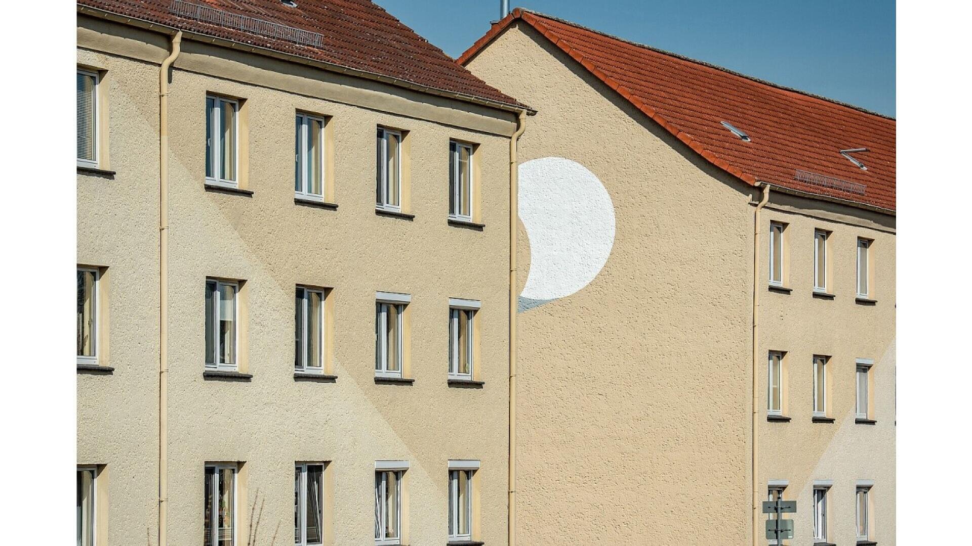 Beige apartment buildings with red-tiled roofs; one building features a painted white circle partially extending onto the wall of the adjacent building, creating an optical illusion in the sunlight.