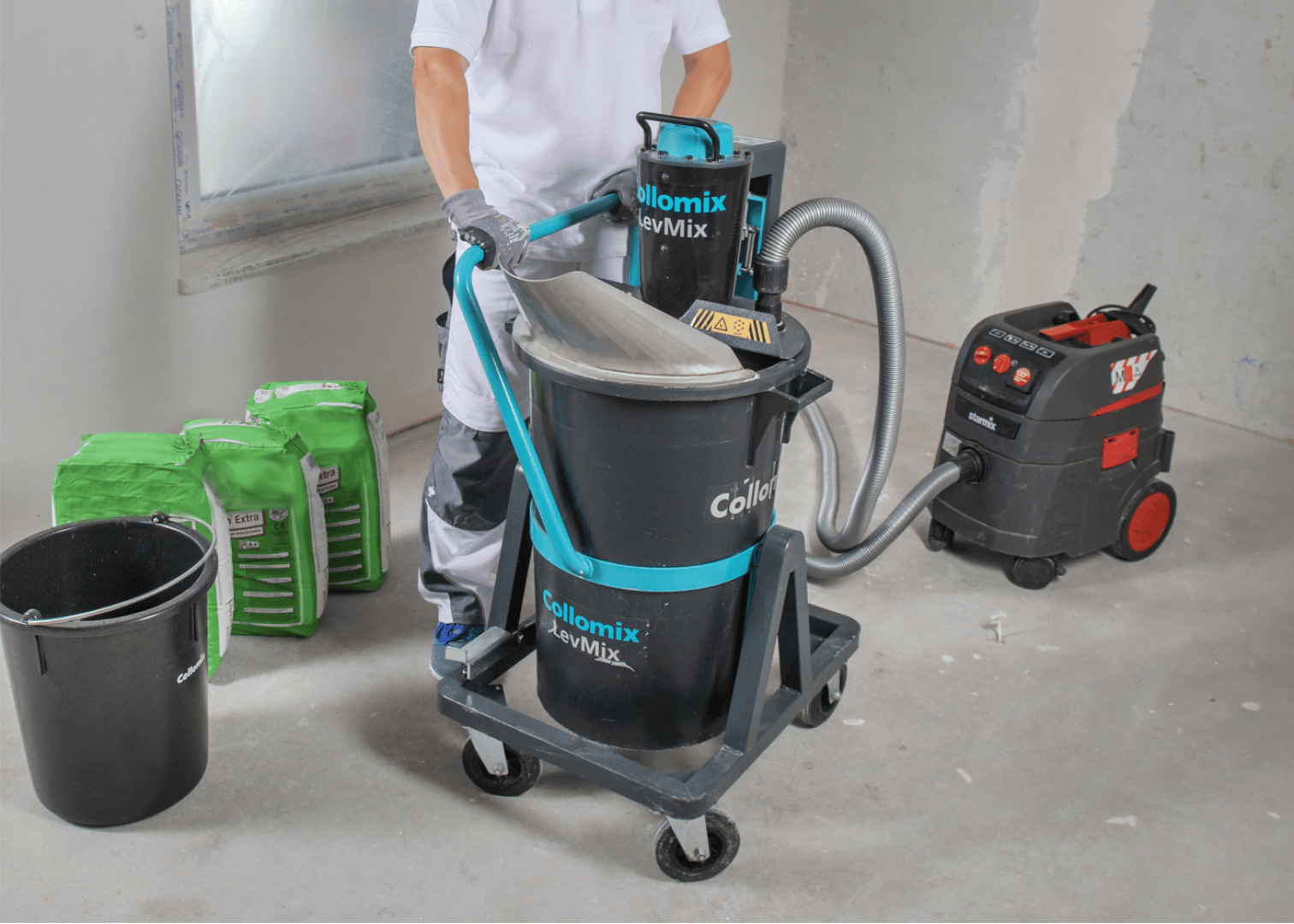 Eine Person in Arbeitskleidung bedient auf einer Baustelle eine Collomix LevMix-Mischmaschine mit Schlauchanschluss. In der Nähe stehen Materialsäcke, ein schwarzer Eimer und eine Vakuummaschine auf einem Betonboden.