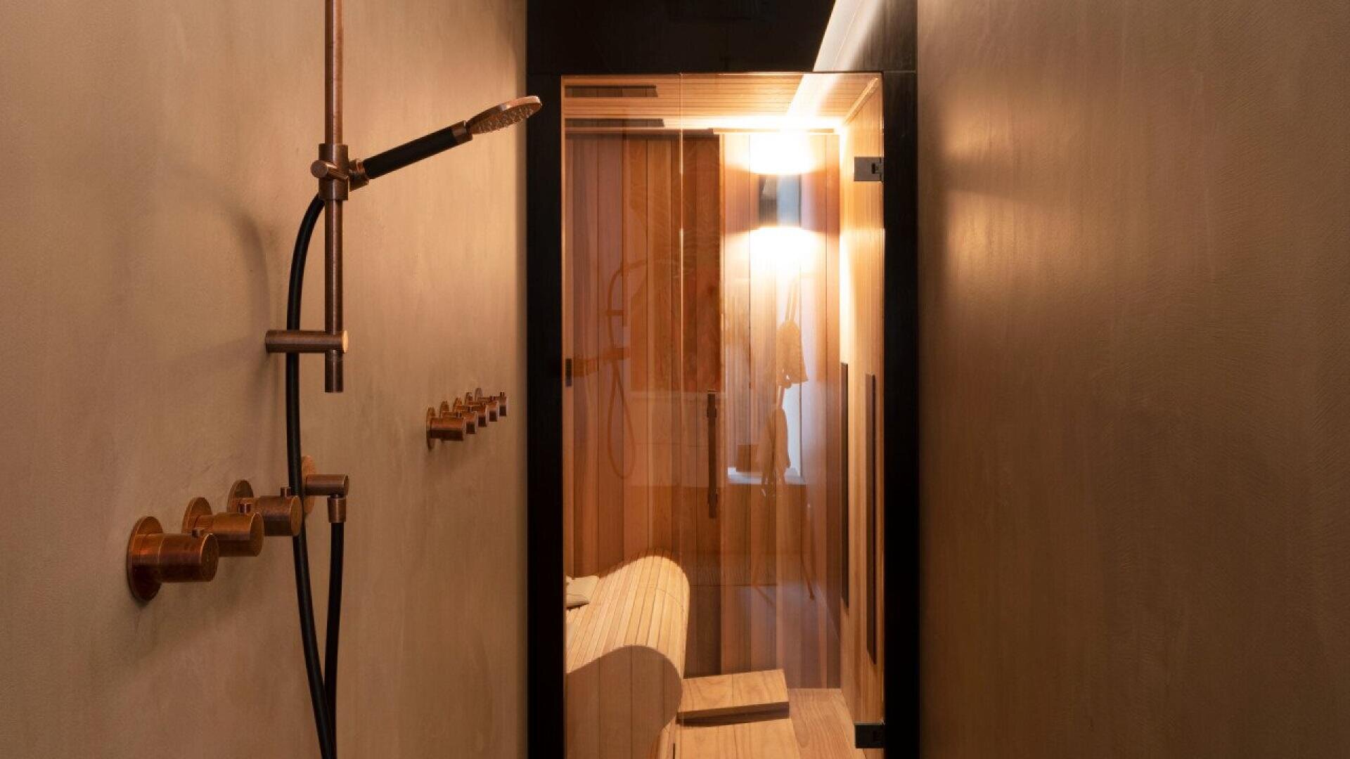 A modern shower with brass fixtures is on the left wall, leading to a glass door that reveals a warm, softly-lit sauna room with wooden benches inside. The overall decor is minimalistic and inviting.