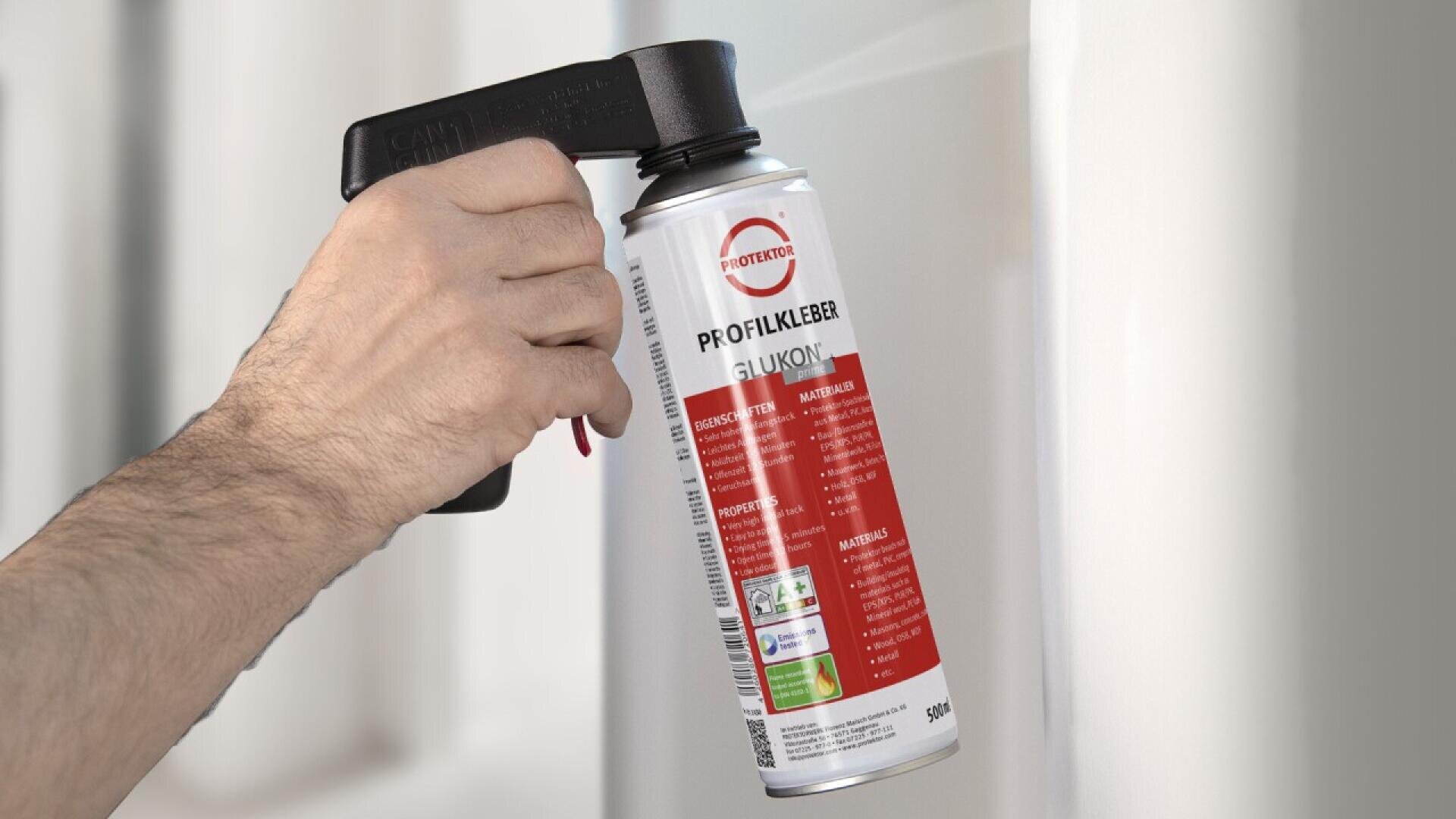 A hand holds a can of spray adhesive equipped with a black spray grip, aiming it towards a white surface. The can has a red and white label with text in German and various symbols.
