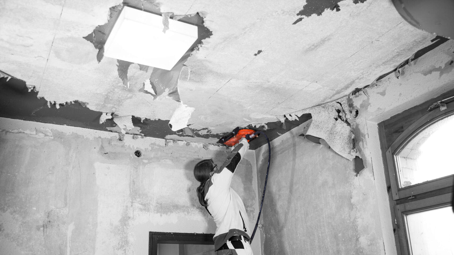 Eine Person entfernt mit einem Elektrowerkzeug abblätternde Farbe oder Tapete von einer beschädigten Decke in einem Raum, der gerade renoviert wird.