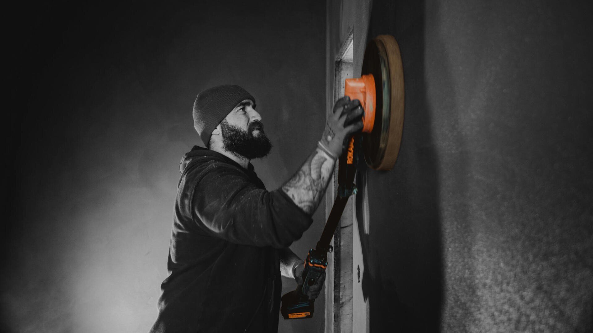 Ein Mann mit Mütze und Kapuzenpullover schleift oder poliert mit einem Elektrowerkzeug eine Wand. Das Bild ist größtenteils schwarz-weiß, das Werkzeug und seine Hand sind orange hervorgehoben.