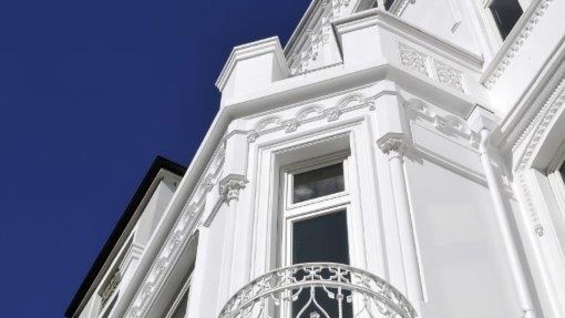 Untersicht eines weißen, reich verzierten Gebäudes im viktorianischen Stil mit dekorativen Stuckarbeiten und einem kleinen Eisenbalkon vor einem klaren, tiefblauen Himmel.