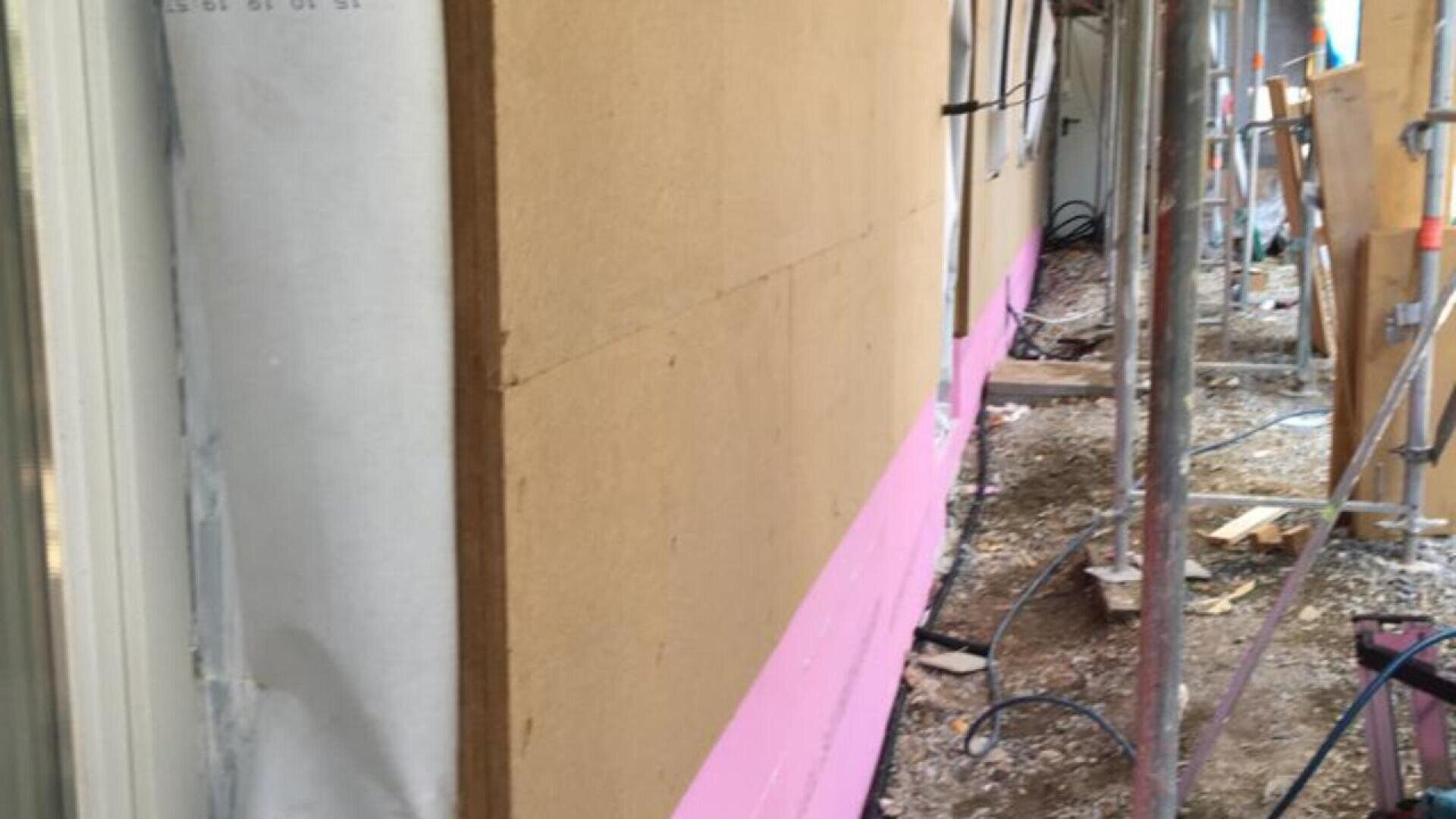 Close-up of a building under construction with beige exterior panels, pink insulation at the bottom, scaffolding alongside, and construction debris on the ground.