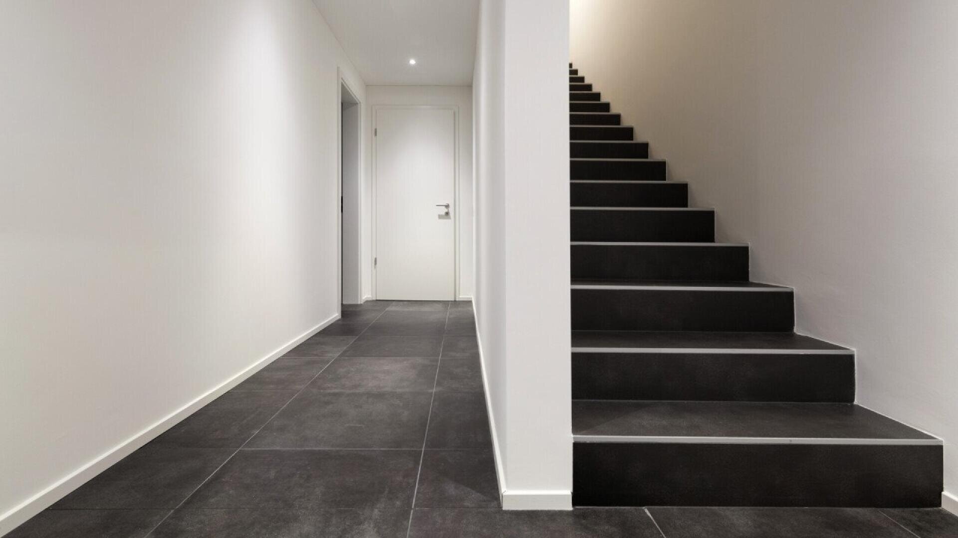 A modern, minimalist hallway with dark tiled floors, white walls, a closed white door at the end, and black stairs with white risers leading upward on the right side. The space is brightly lit.
