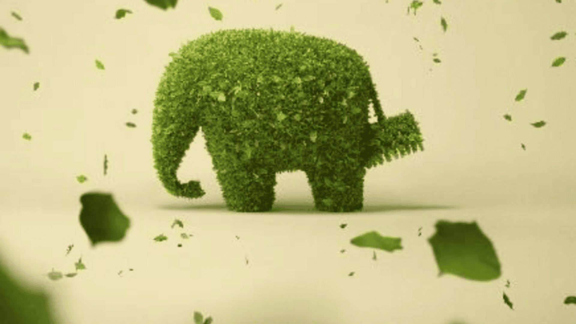 A sculpture of an elephant made entirely of green leaves or grass stands on a light background, with green leaves swirling around it in the air.