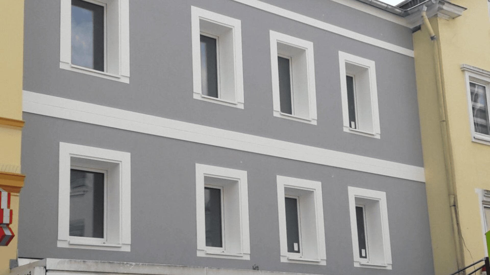 Ein graues Gebäude mit weißen Verzierungen verfügt über zwei Reihen mit je drei rechteckigen Fenstern, die jeweils von weißen Rahmen umgeben sind. Die angrenzenden Gebäude sind gelb gestrichen.