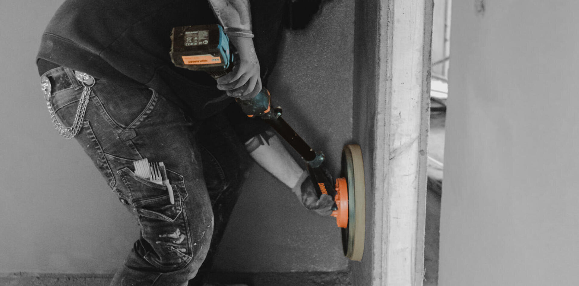Eine Person in Jeans mit Werkzeugen in den Taschen glättet mit einem elektrischen Schleifgerät eine Wand. Das Bild ist größtenteils schwarz-weiß, mit Ausnahme des Schleifgeräts, das farblich hervorgehoben ist.