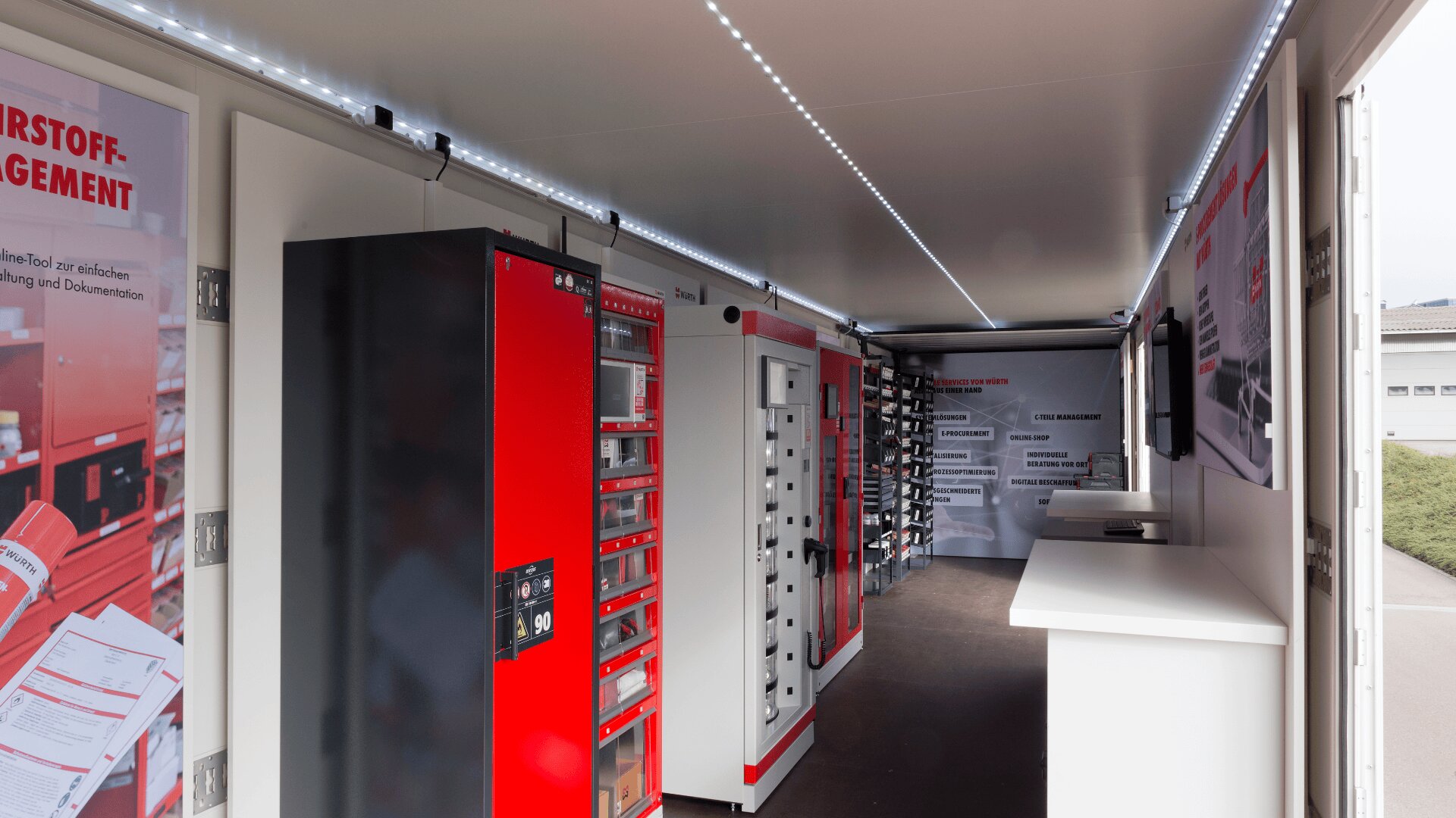 Innenraum eines modernen Industrieanhängers mit rot-schwarzen Geräteschränken, Postern an den Wänden, einer weißen Theke und LED-Lichtbändern an der Decke. Ein Fenster auf der rechten Seite sorgt für Tageslicht.