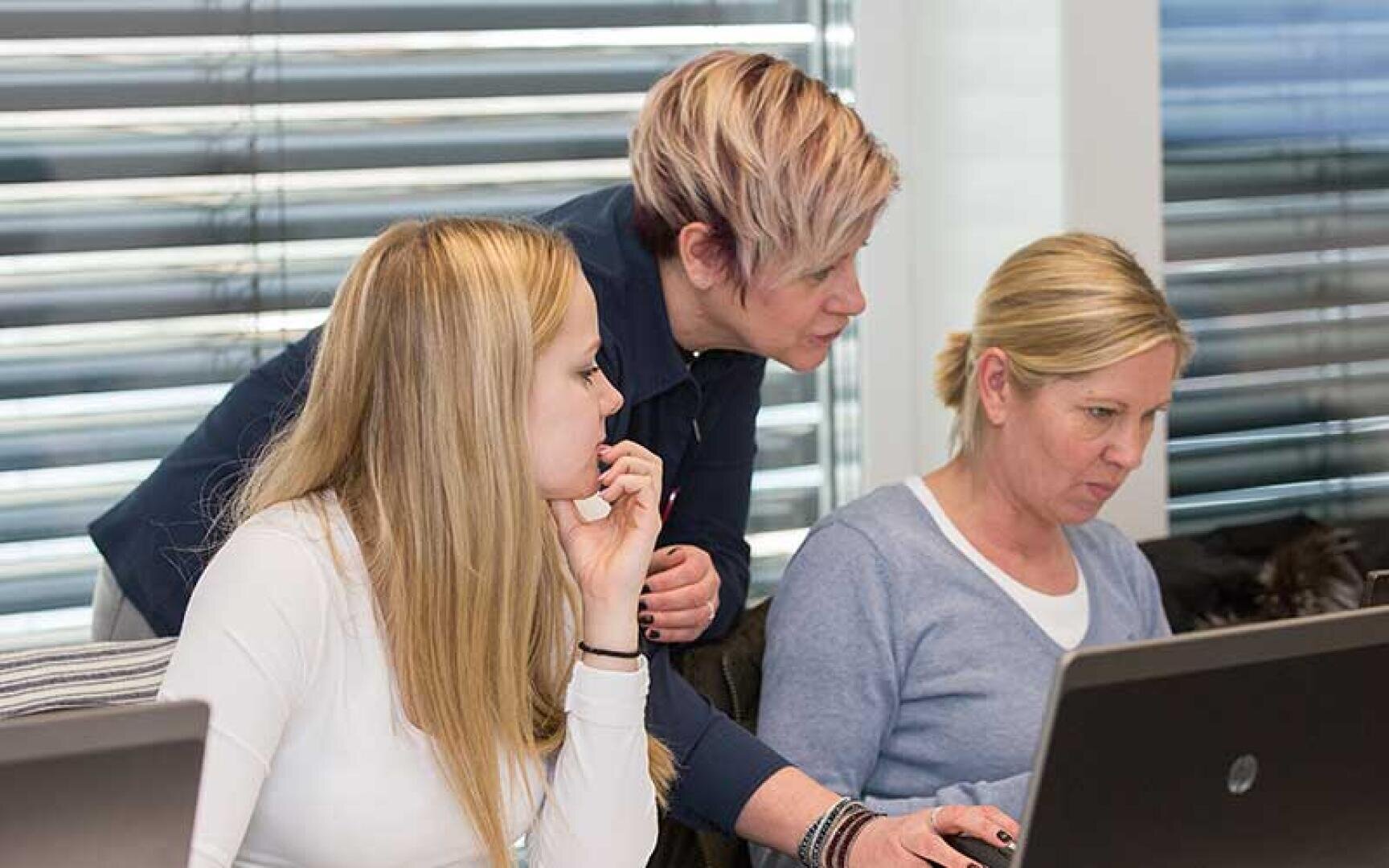 Drei Frauen arbeiten konzentriert an ihren Laptops in einem Büro. Eine steht auf und beugt sich vor, um den beiden sitzenden Frauen zu helfen, die konzentriert an ihren Computern arbeiten. Horizontale Jalousien verdecken die Fenster hinter ihnen.