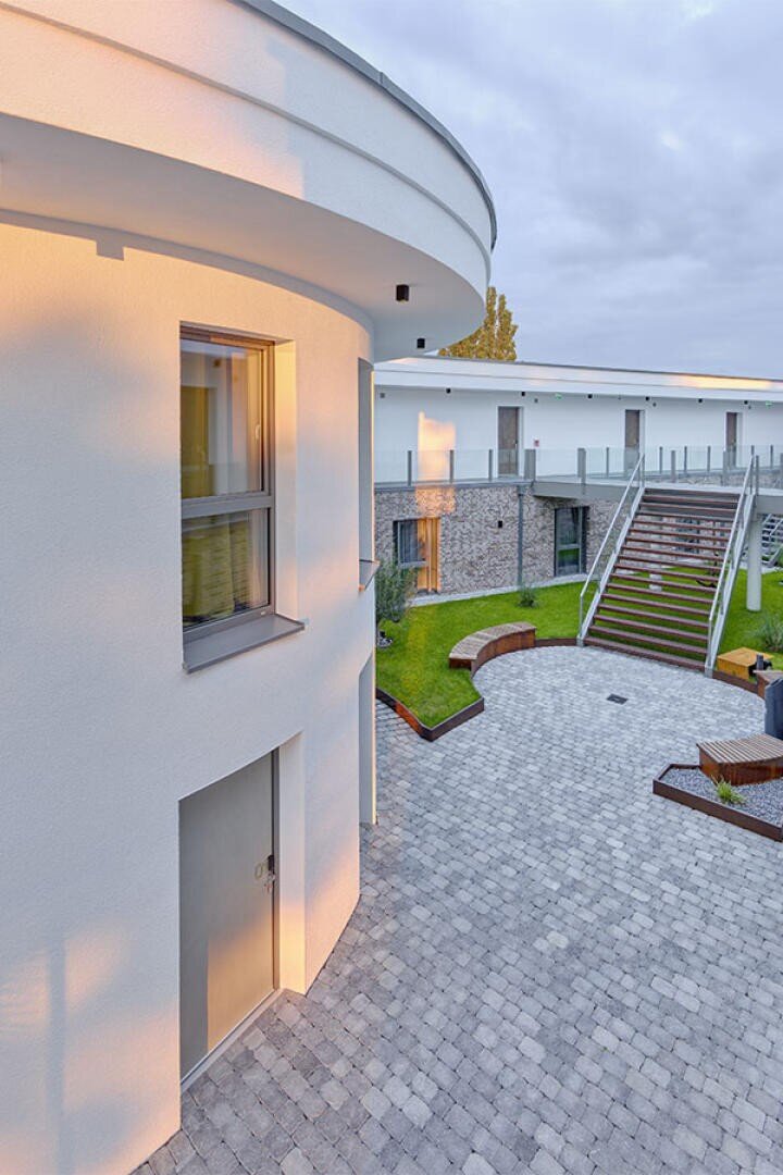 Moderner Innenhof mit einem geschwungenen weißen Gebäude, großen Fenstern und einem gepflasterten Gehweg. Es gibt Bänke, grünes Gras, Pflanzen und eine Treppe mit einem Glasgeländer, die zu einem Gehweg im oberen Stockwerk führt.