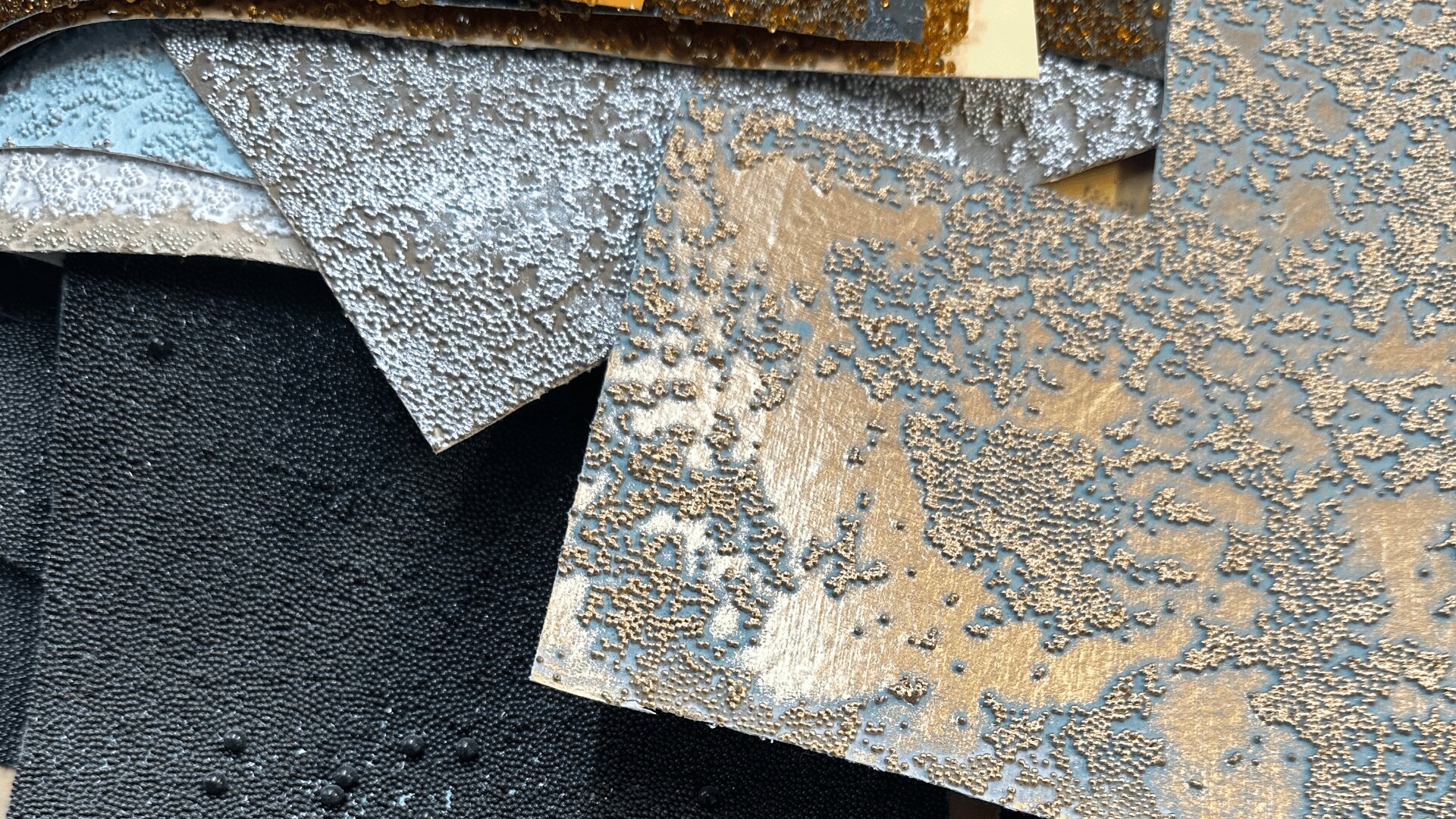 Close-up of several overlapping textured sheets in shades of black, gray, and gold, featuring rough, bubbly, and uneven surfaces creating a tactile, abstract pattern.