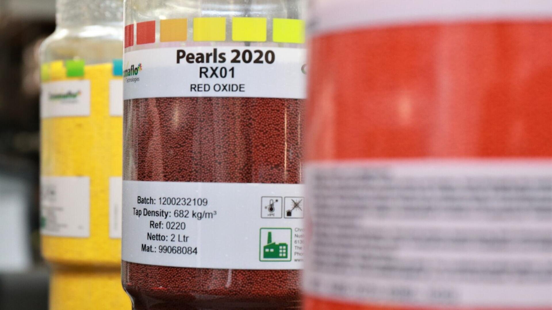 Close-up of containers filled with colored granules, focusing on a label reading Pearls 2020 RX01 Red Oxide. Other containers with yellow and red contents are visible in the background and foreground.