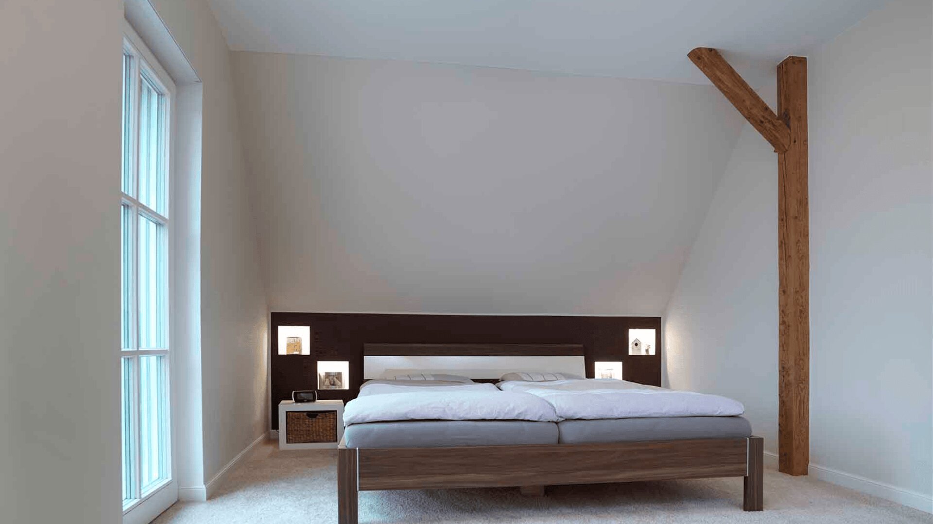 Minimalistisches Schlafzimmer mit Doppelbett, weißen Wänden, großem Fenster auf der linken Seite, eingebautem Regal mit Beleuchtung am Kopfteil, Nachttisch und freiliegendem Holzstützbalken in der Ecke.