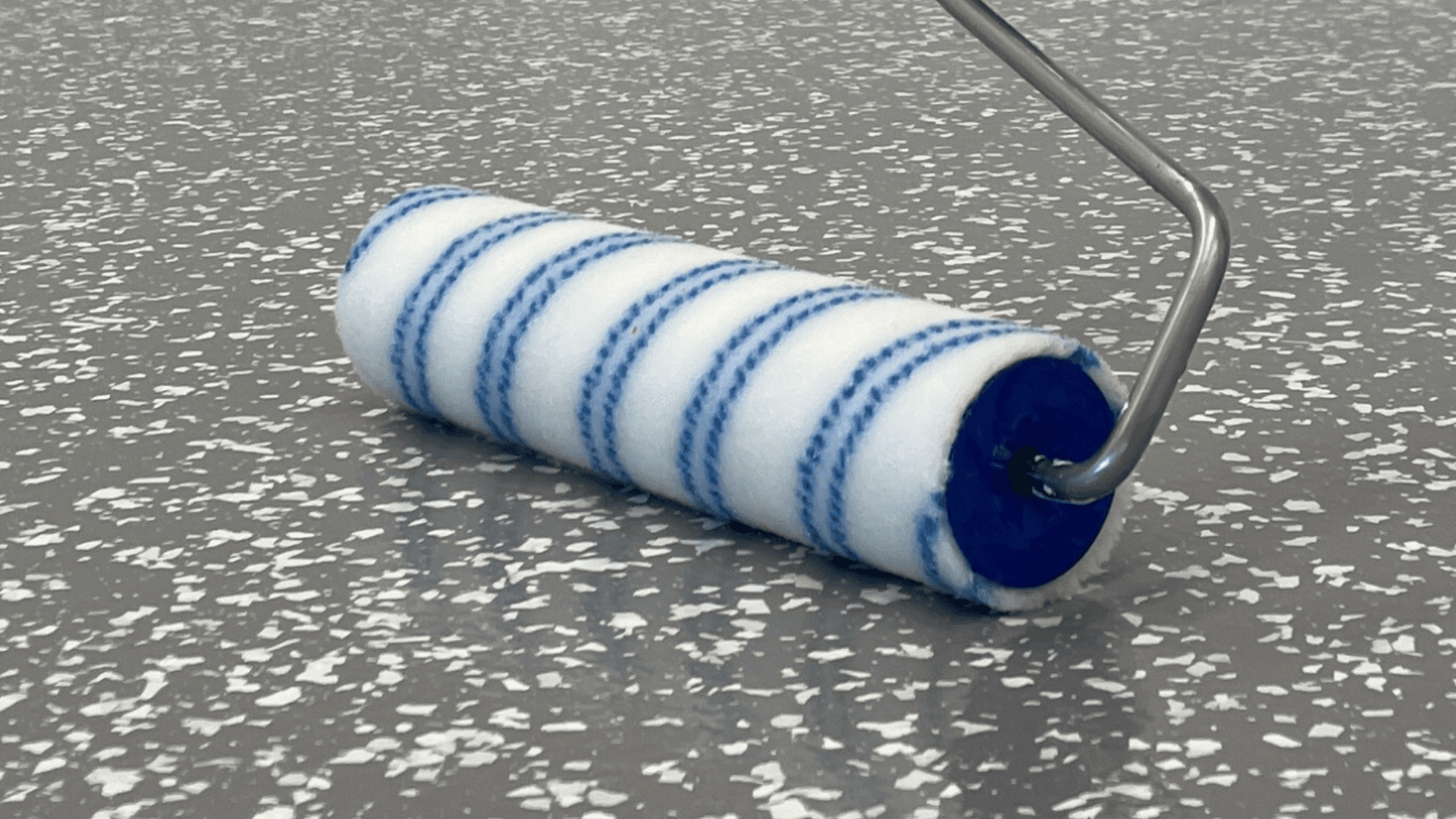 Eine Nahaufnahme einer Farbrolle mit weißen und blauen Streifen auf einer grauen, gesprenkelten Oberfläche, die einem gestrichenen oder mit Epoxidharz beschichteten Boden ähnelt.