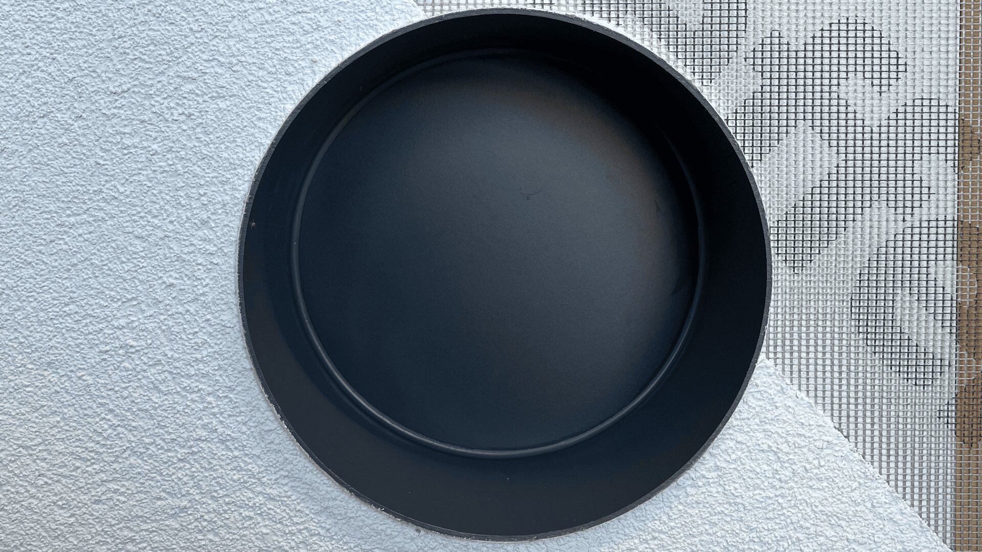 Ein schwarzer, runder, leerer Teller steht auf einer strukturierten weißen Oberfläche, in der oberen rechten Ecke ist ein Teil einer grauen Matte mit geometrischem Muster sichtbar.