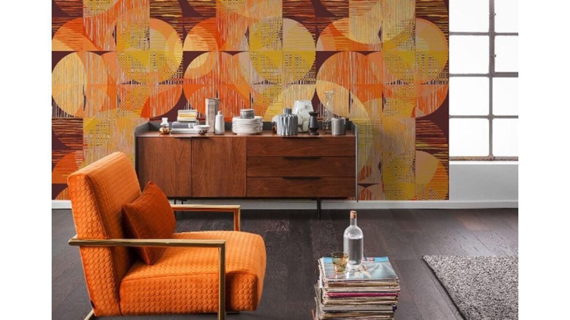 A modern living room with an orange armchair, a small glass table with stacked books and a bottle, a wooden sideboard with various dishes, and a bold geometric orange and yellow accent wall. Large window frames brighten the space.
