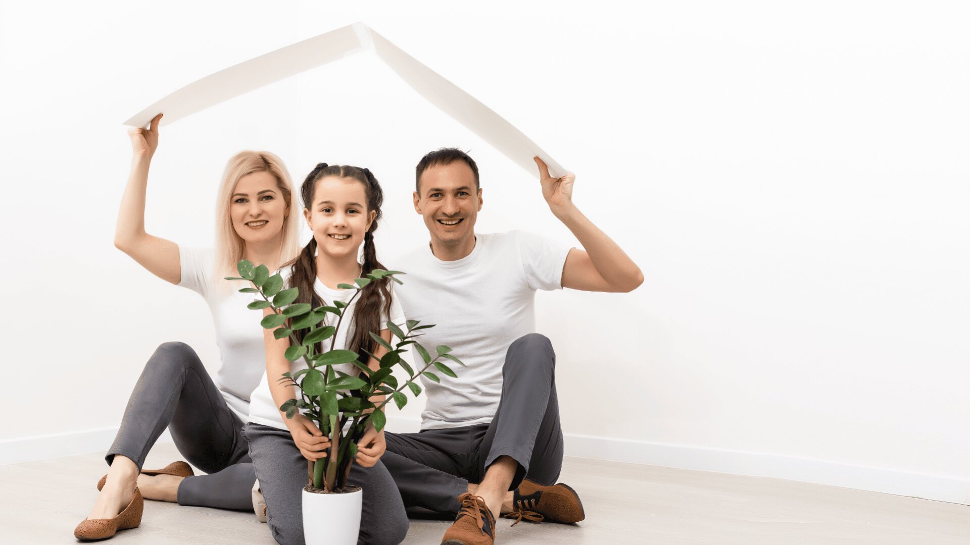 Eine lächelnde dreiköpfige Familie sitzt auf dem Boden vor einer weißen Wand und hält ein Papierdach über ihren Köpfen. Zwischen zwei Erwachsenen sitzt ein junges Mädchen, vor ihnen steht eine grüne Topfpflanze.