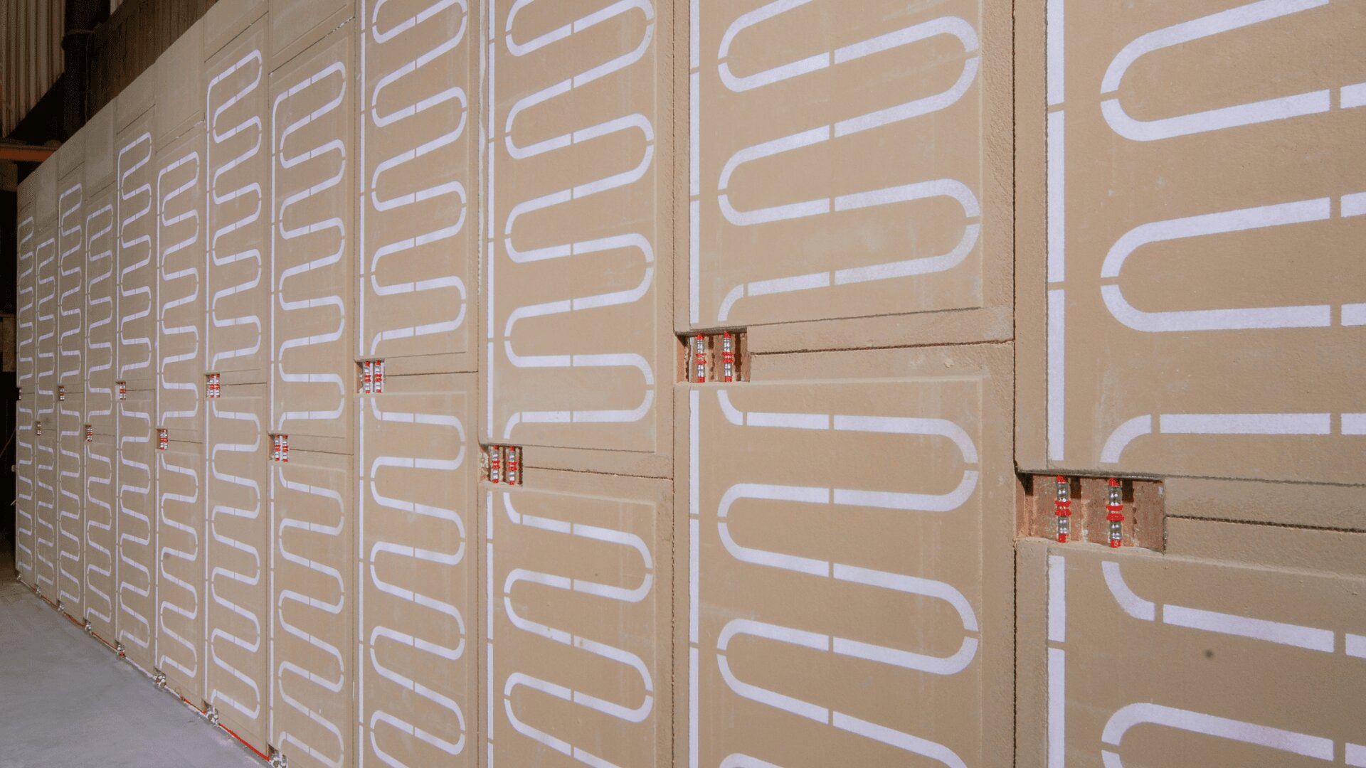 Eine Wand, die mit großen beigen Dämmplatten verkleidet ist und in die weiße, gewundene Heizungsrohre in einem sich wiederholenden Muster eingebettet sind, möglicherweise für die Installation einer Strahlungsheizung.