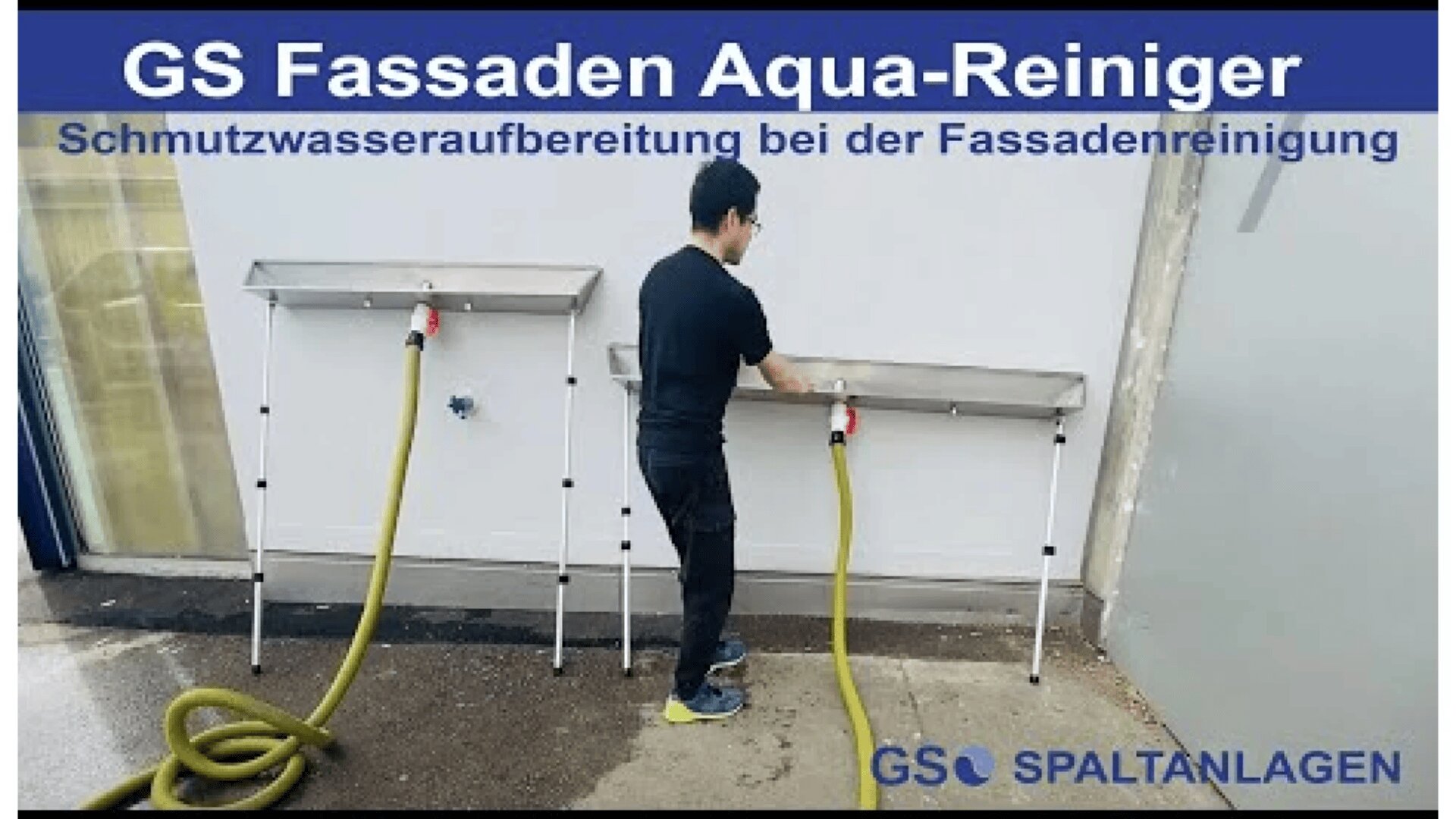 Ein Mann benutzt einen Schlauch, der an einer Metallreinigungsstation mit der Aufschrift „GS Fassaden Aqua-Reiniger“ an einer weißen Wand befestigt ist. Auf dem Text steht „Abwasseraufbereitung zur Fassadenreinigung“.