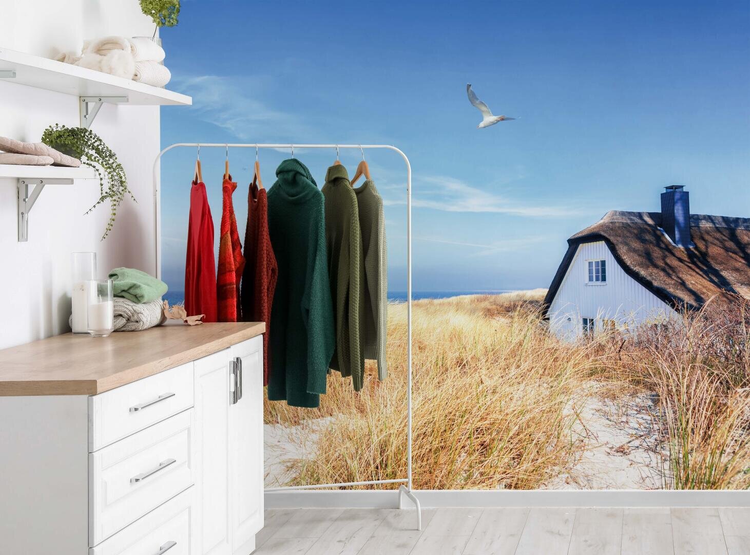 Eine helle Waschküche mit einer Arbeitsplatte, Regalen, gefalteten Handtüchern, einem Kleiderständer mit bunten Pullovern und einem Wandgemälde, das ein Strandhaus, Sanddünen und eine fliegende Möwe unter einem blauen Himmel zeigt.