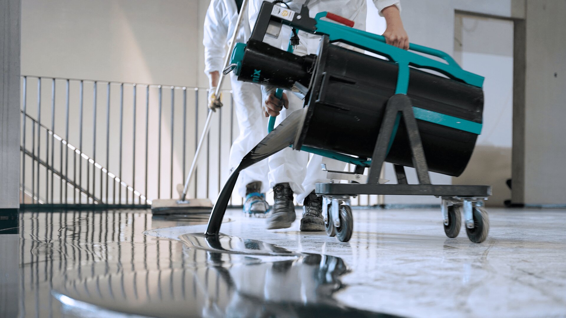 Zwei Arbeiter in Schutzanzügen tragen mit einer großen Maschine auf Rädern eine flüssige Beschichtung auf einen Boden auf, während einer mit einem Werkzeug die Beschichtung gleichmäßig verteilt. Die Oberfläche erscheint glatt und spiegelnd.