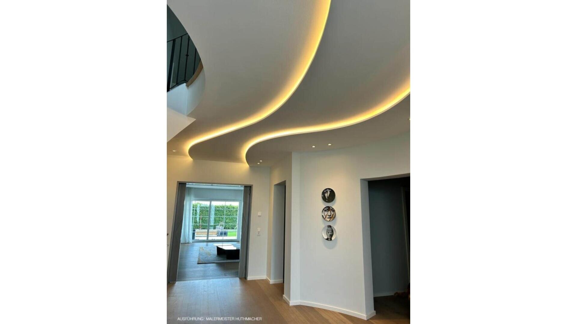 A modern interior with a curved ceiling featuring recessed LED strip lighting, white walls, wooden floors, and three decorative plates on the wall. Large windows in an adjacent room let in natural light.