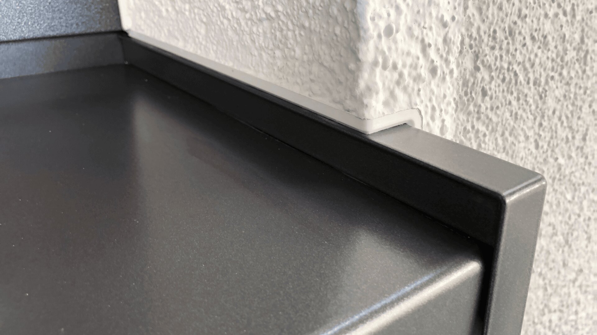Nahaufnahme einer metallischen Oberfläche mit erhabener Kante, die genau an eine strukturierte, weiße Wandecke passt. Die Oberflächen wirken klar und modern, mit scharfen Linien und glatten Oberflächen.