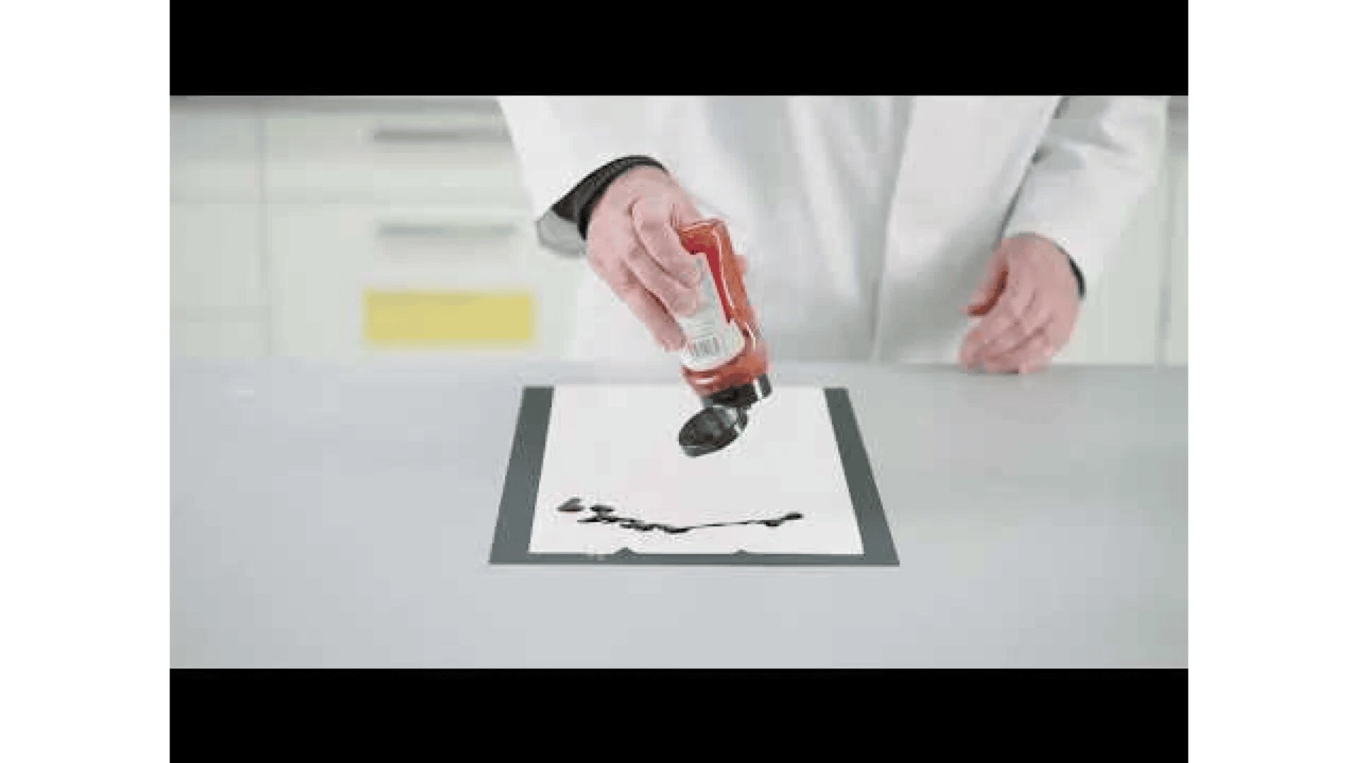 Eine Person im weißen Laborkittel drückt eine Flasche braune Soße auf ein weißes Blatt Papier auf einem grauen Tisch in einer Laborumgebung.