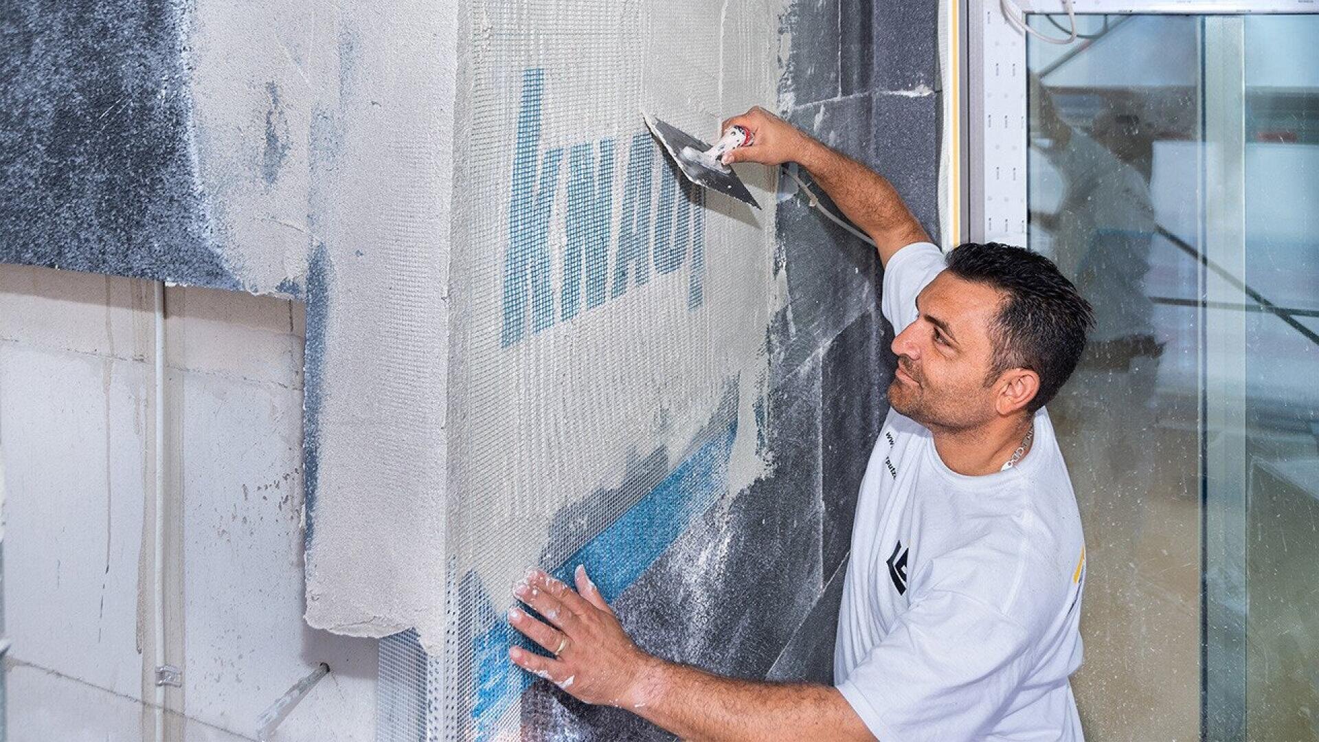 A man in a white shirt smooths plaster onto a wall with a trowel, reinforcing it with mesh material in a construction or renovation setting.