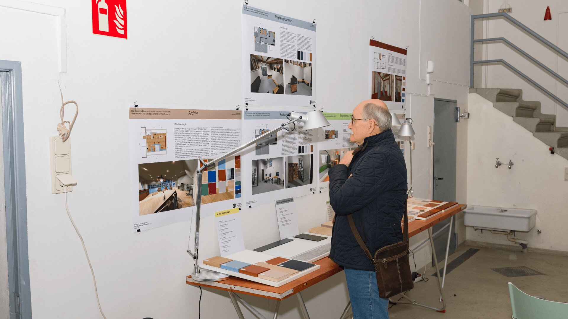 Eine Person steht in einem gut beleuchteten Innenraum vor einer Schautafel mit Architekturplänen, Fotos und Farbmustern. Sie scheint die Materialien aufmerksam zu studieren.