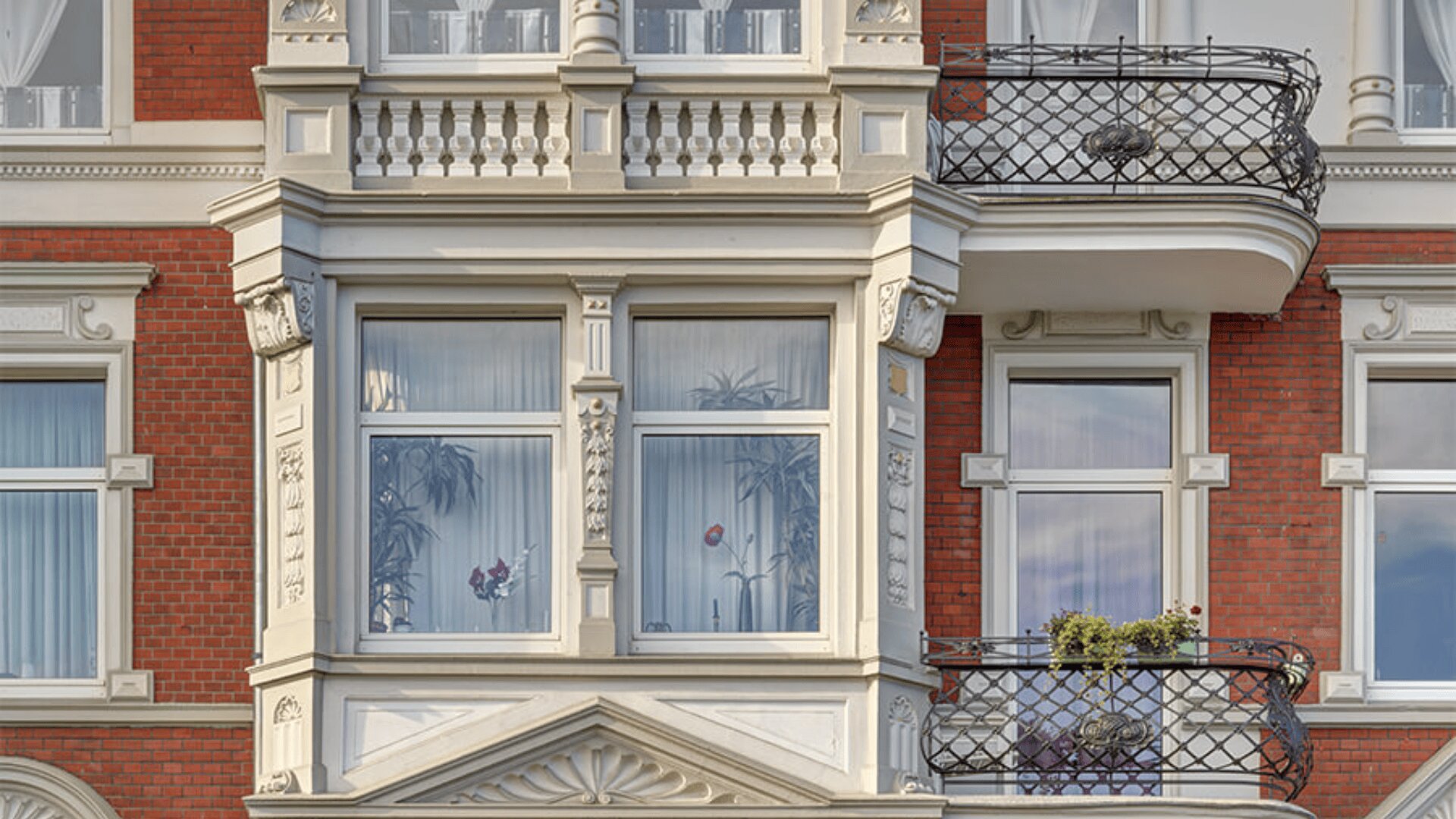 Nahaufnahme einer kunstvollen Gebäudefassade mit großen Fenstern, weißen Zierleisten, roten Backsteinmauern und schmiedeeisernen Balkonen, teilweise mit Topfpflanzen und Blumen. Durch die Fenster sind transparente Vorhänge und Zimmerpflanzen sichtbar.