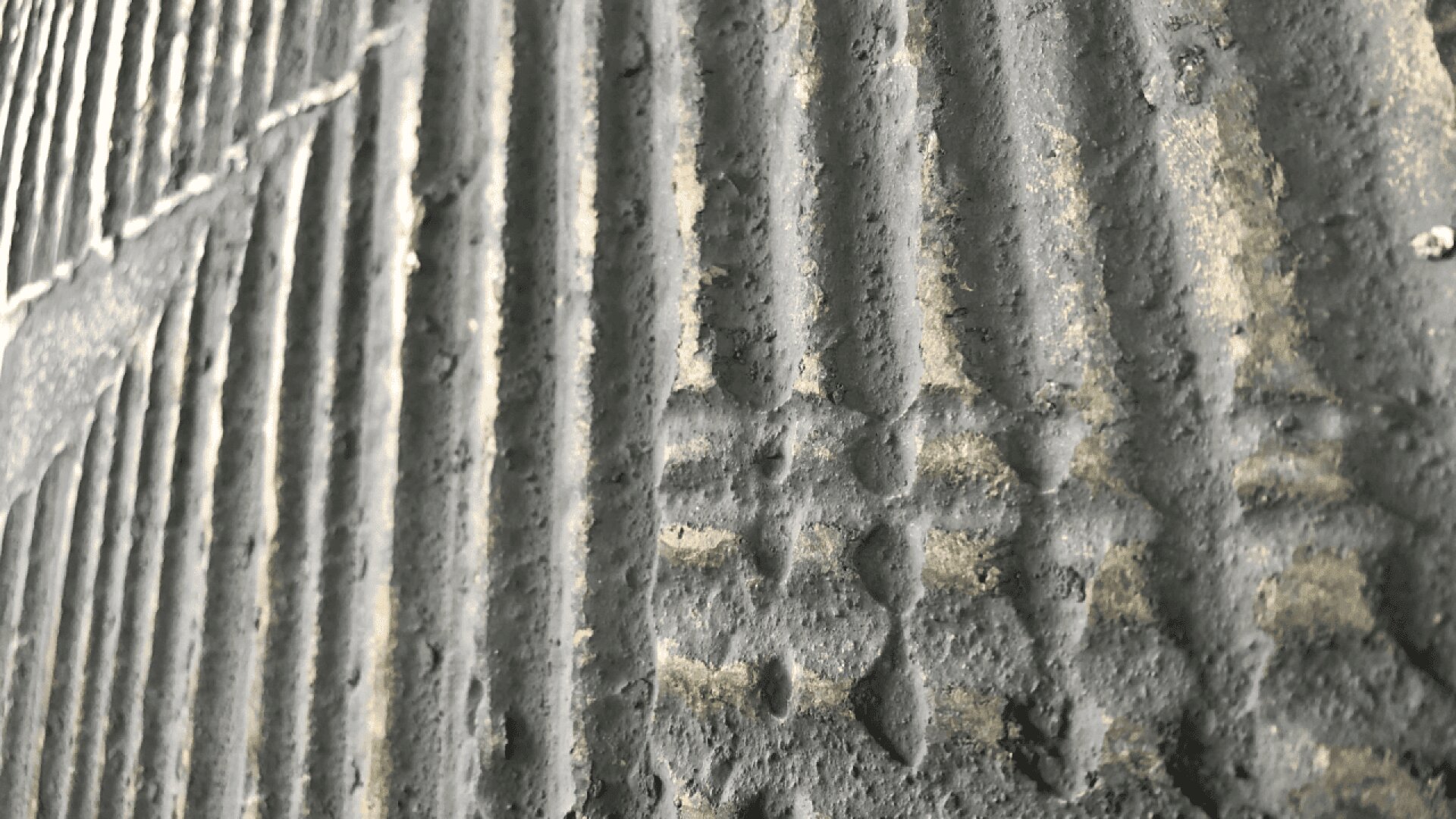 Close-up of a textured, grooved concrete or stone wall with diagonal and vertical lines; the surface appears rough and uneven with patches of lighter and darker gray.